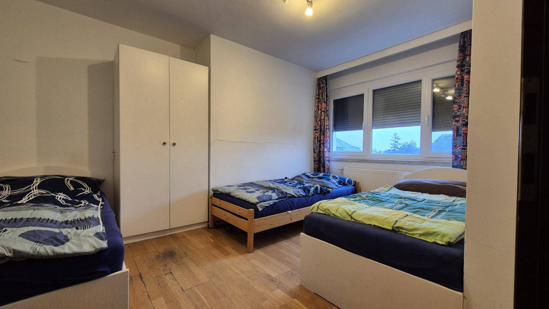 Schlafzimmer mit drei Einzelbetten, Holzboden und einem großen Fenster mit Rollläden.