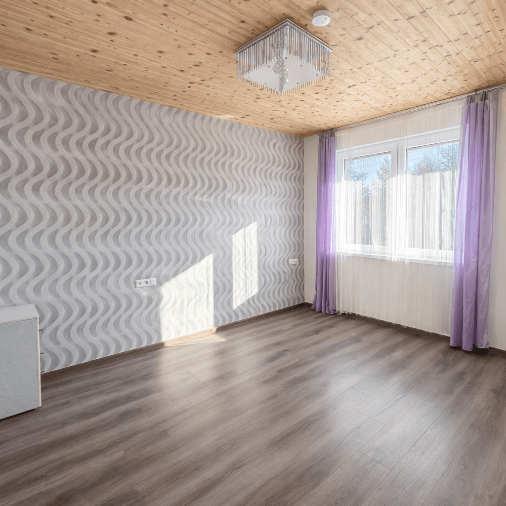 Helles Zimmer mit Holzbalkendecke, gemusterter Tapete und Laminatboden, durchflutet von Sonnenlicht.