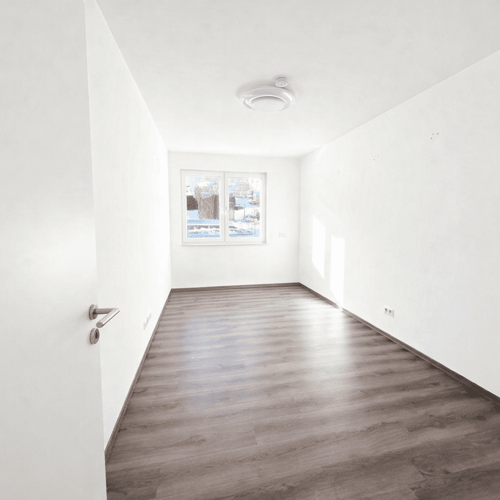 Leerer Raum mit Laminatboden, weißen Wänden und einem Fenster, das viel Tageslicht spendet.