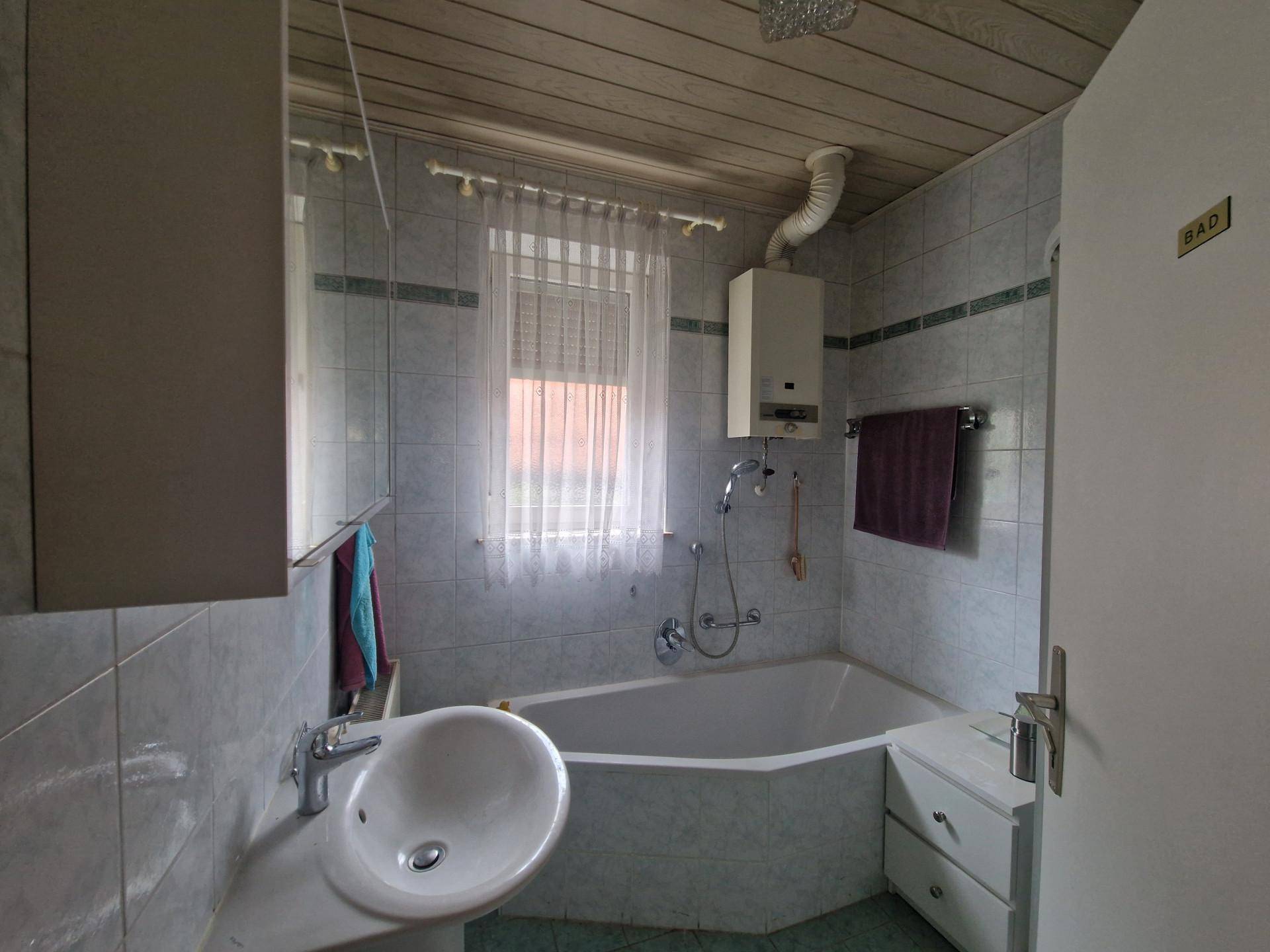 Badezimmer mit Badewanne, Waschbecken und Fenster, ausgestattet mit hellen Fliesen und Holzdecke.