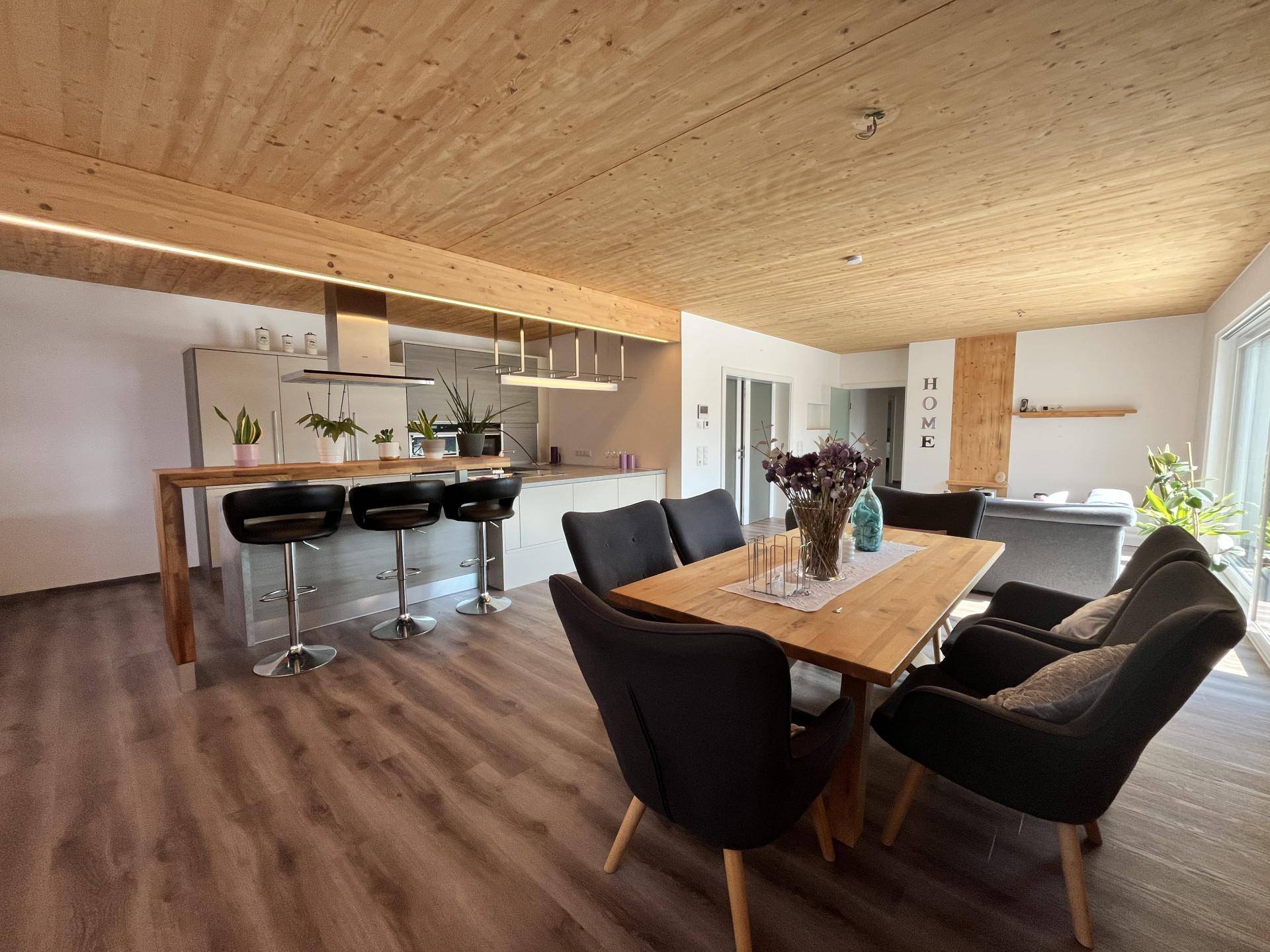 Offener Wohn-Essbereich mit moderner Küche, Holzbalkendecke und dunklem Laminatboden.