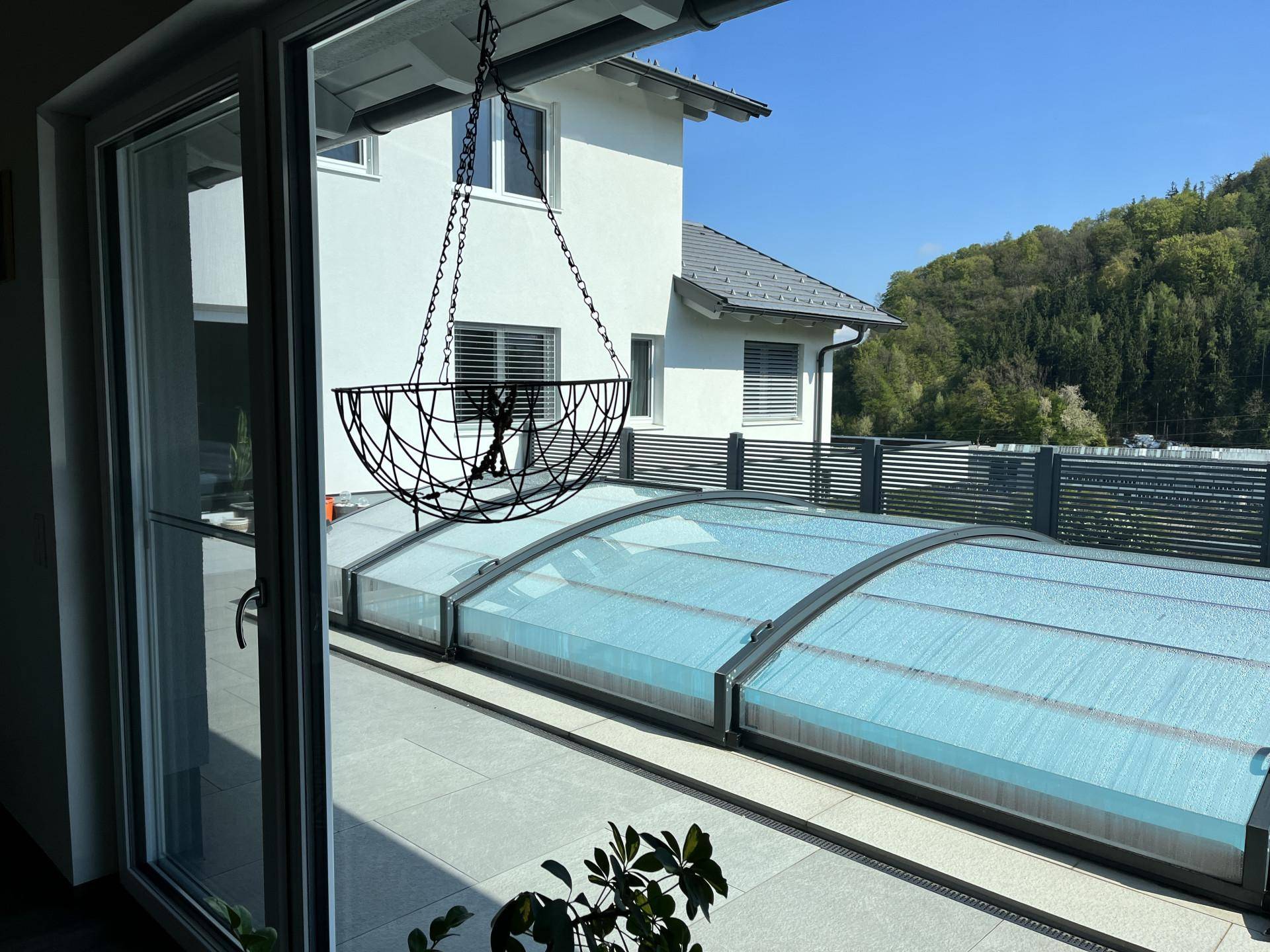 Blick von innen auf die sonnige Terrasse mit überdachtem Pool und umliegender Natur.