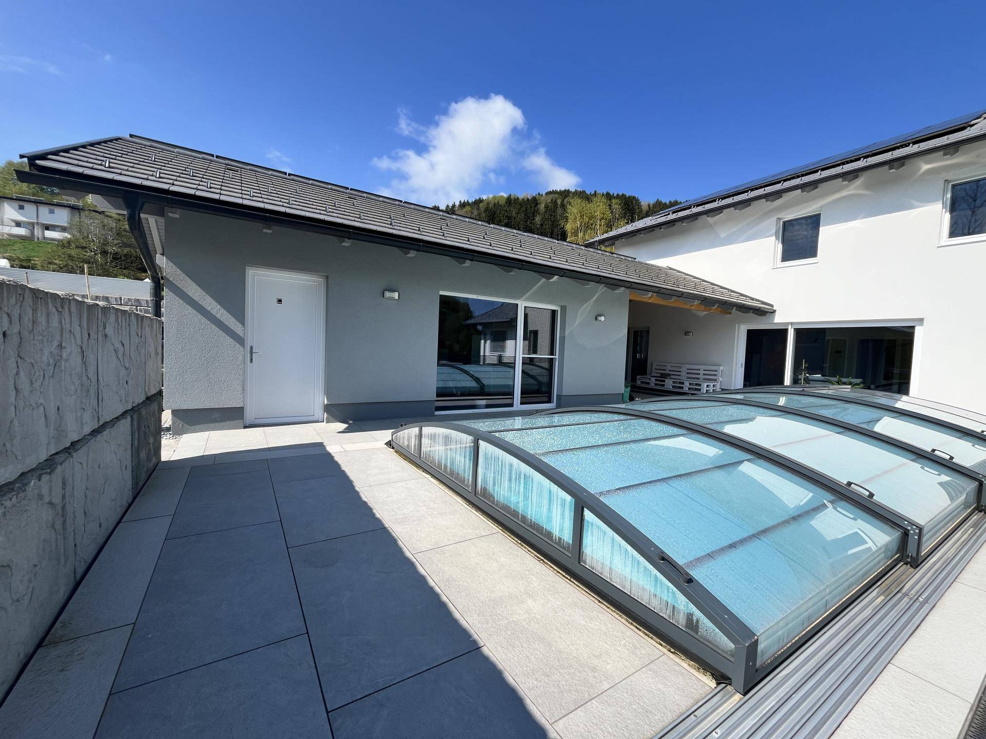 Außenansicht des Hauses mit großzügiger Terrasse und überdachtem Swimmingpool unter blauem Himmel.