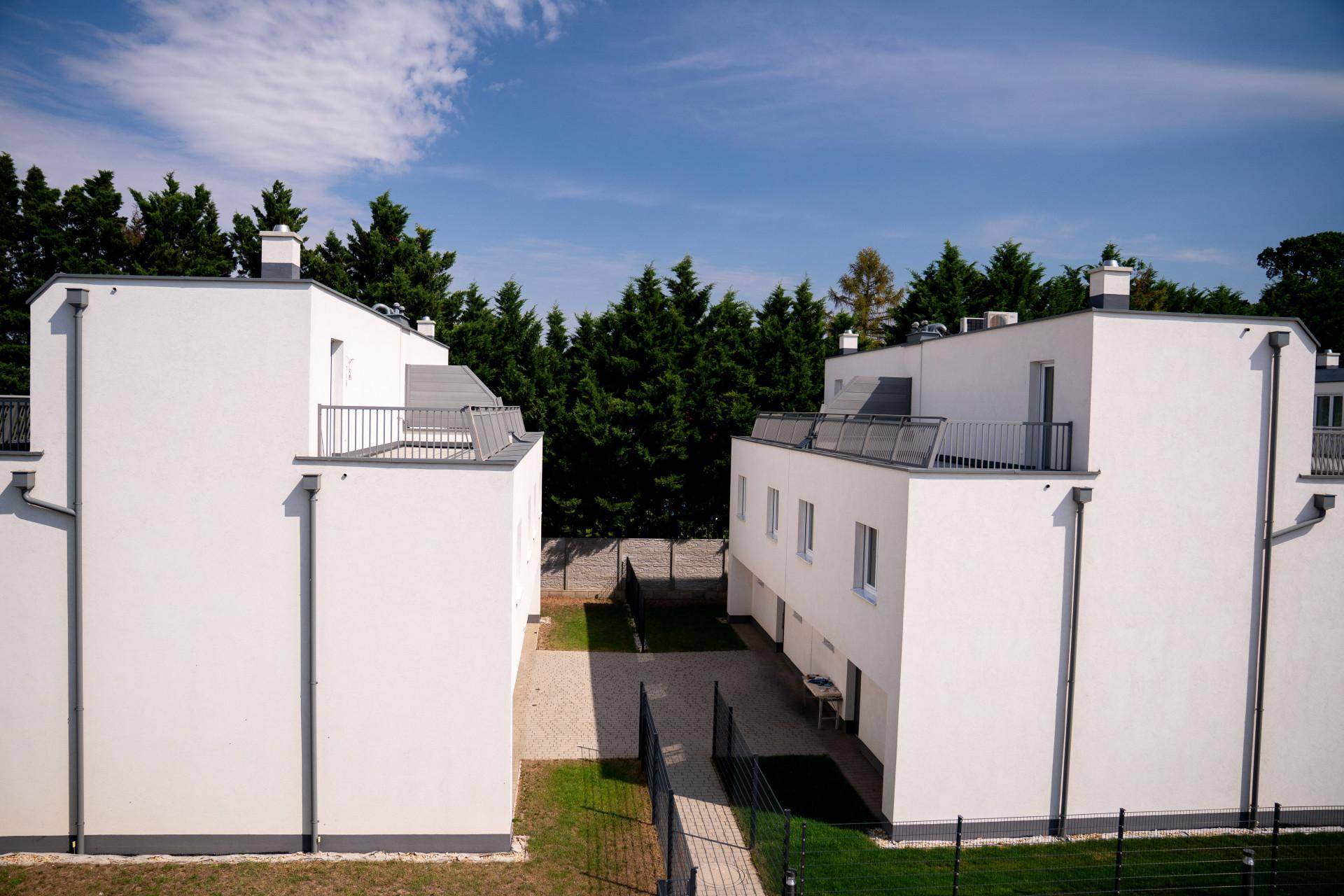 Zwei moderne weiße Gebäude mit Flachdächern und einem begrünten Innenhof unter blauem Himmel.