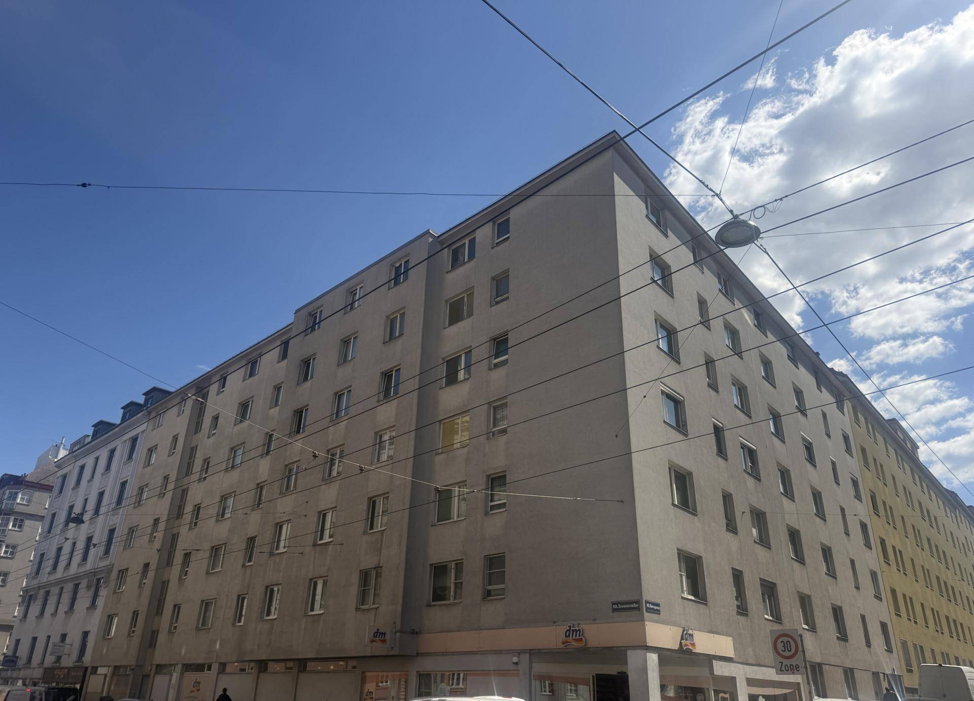 Großes Wohngebäude mit grauer Fassade und Geschäften im Erdgeschoss an einer Stadtstraße.