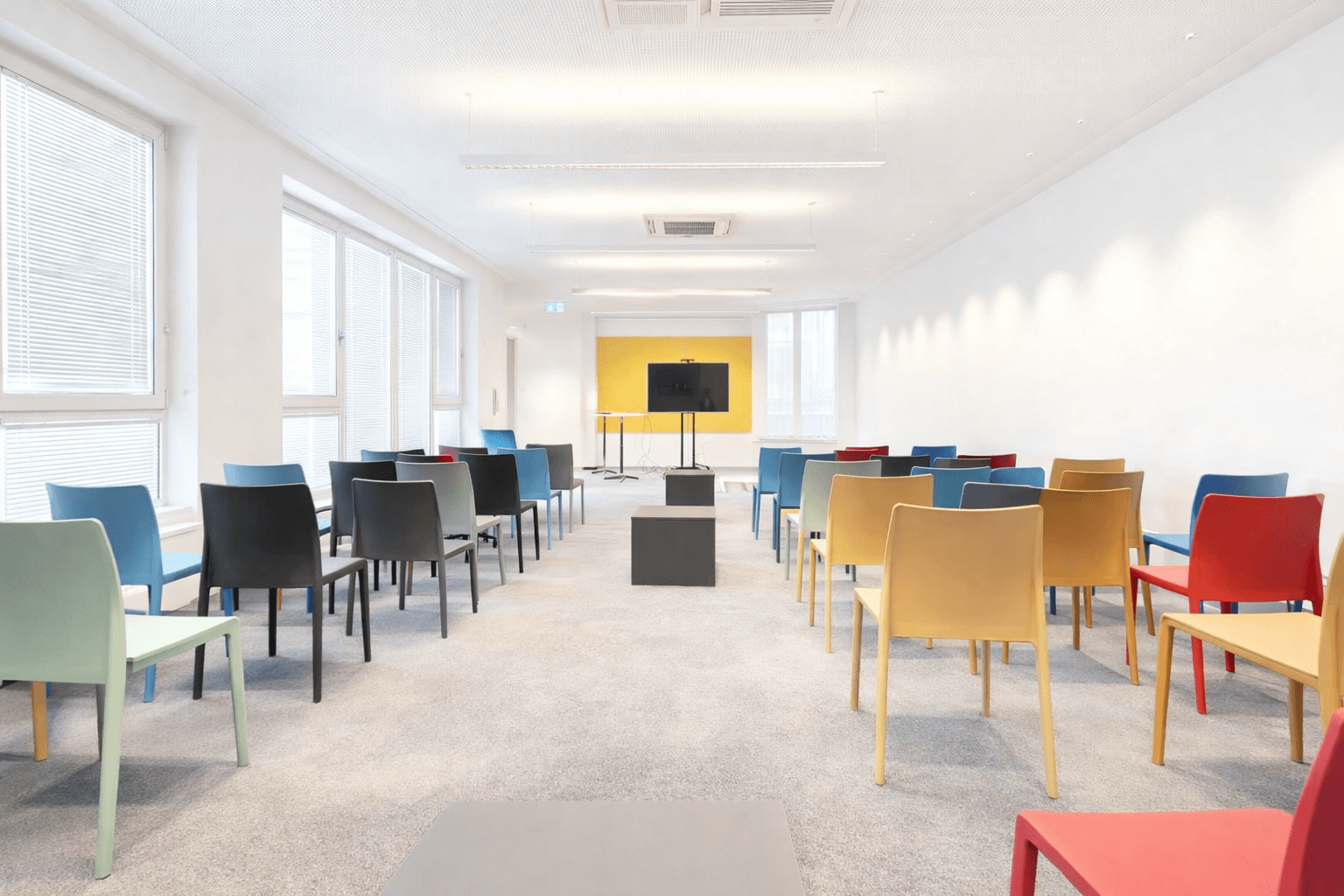 Heller Konferenzraum mit bunten Stühlen, Teppichboden und einem Bildschirm an der gelben Akzentwand.