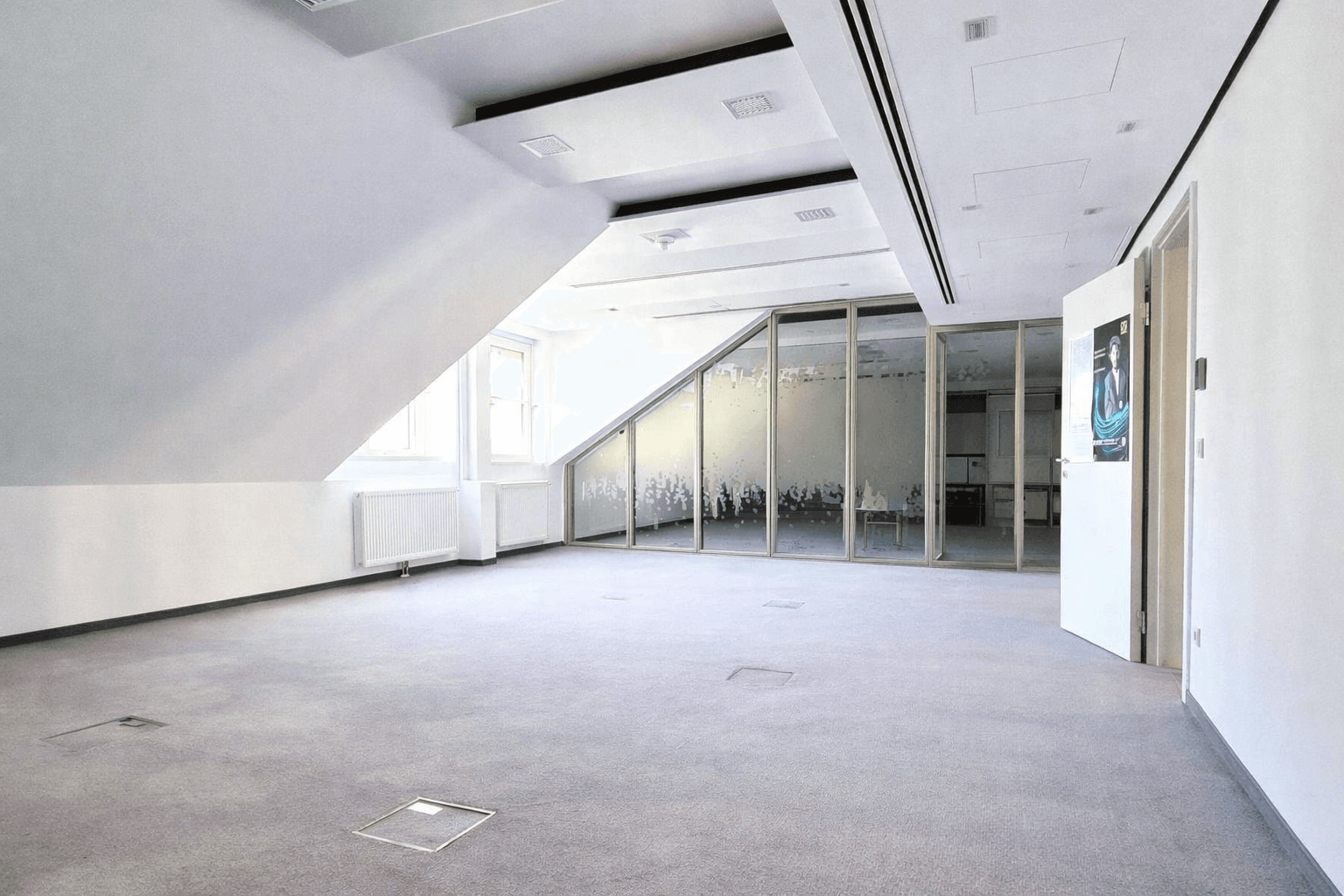 Helles, leeres Büro mit Teppichboden, schrägen Wänden und Glaswänden zu angrenzenden Bereichen.