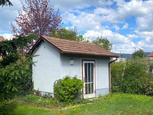 Kleines Gartenhaus mit Ziegeldach, umgeben von Bäumen und Sträuchern im Grünen.