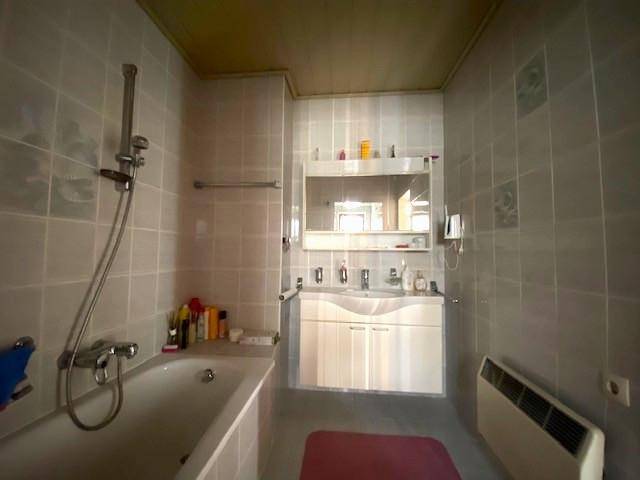 Badezimmer mit hellgrauen Fliesen, Badewanne mit Duschfunktion und einem Waschtischunterschrank.