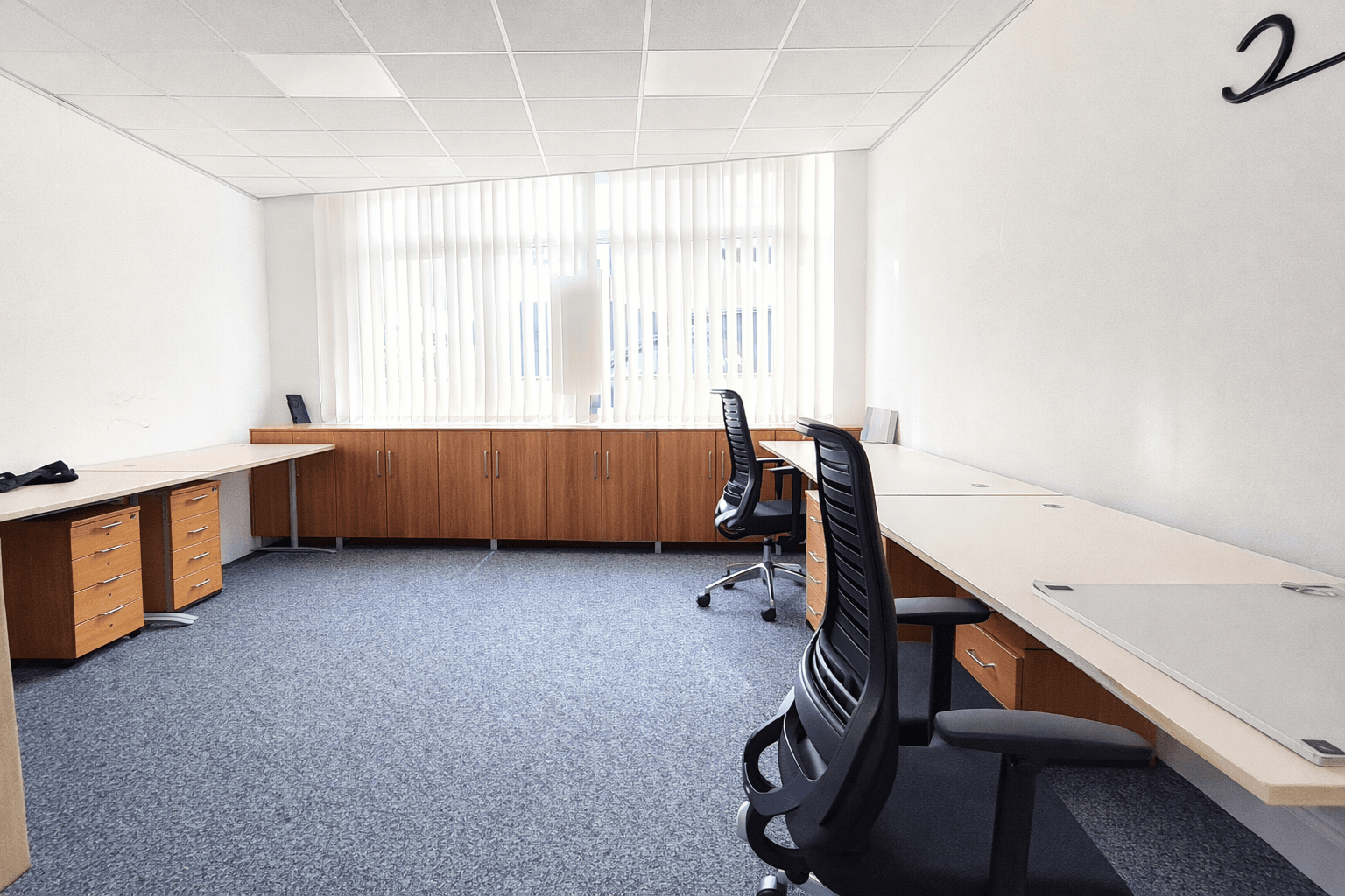 Großes Büro mit mehreren Arbeitsplätzen, Teppichboden und langen Fenstern mit Jalousien.