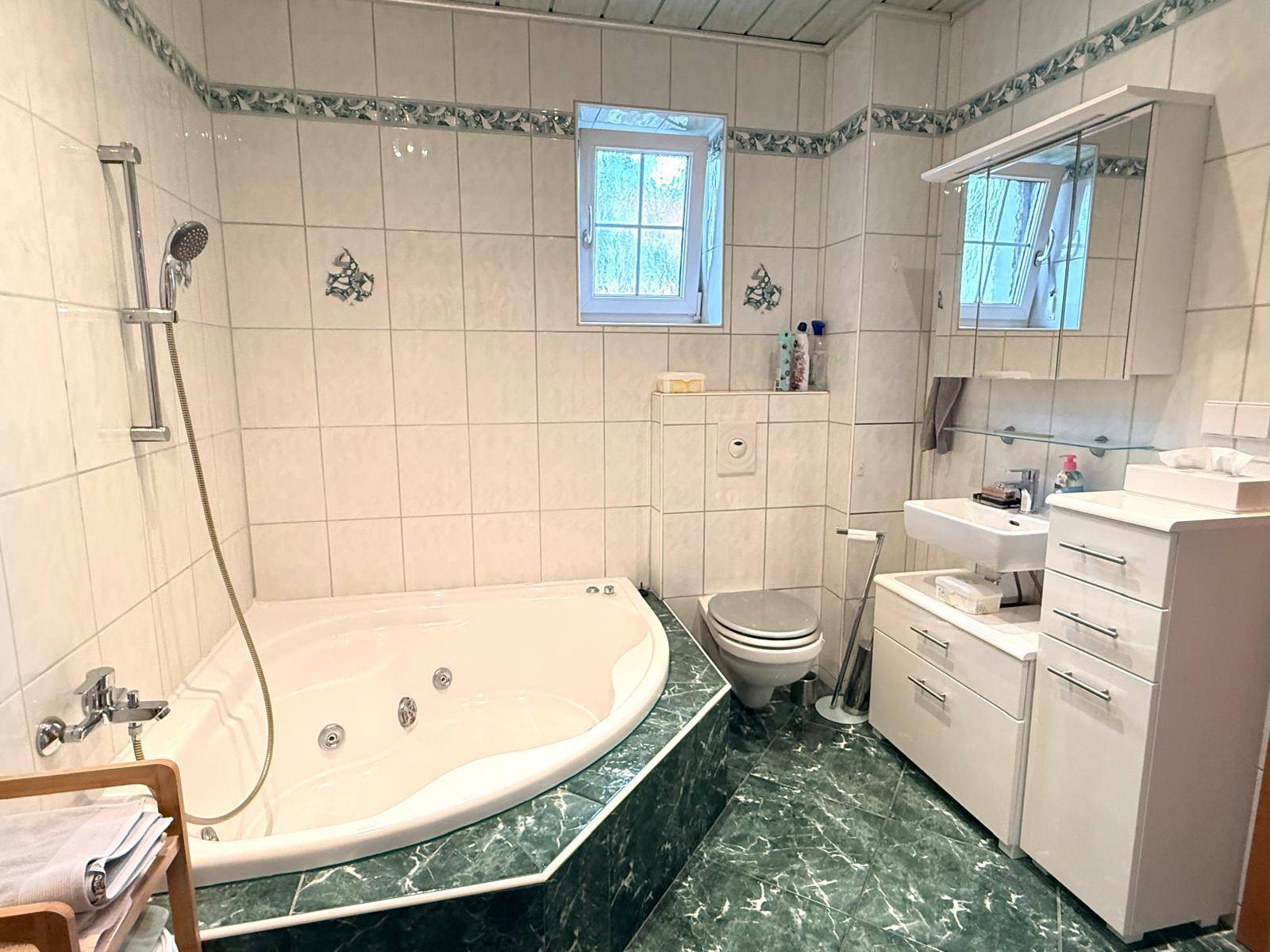 Badezimmer mit Eckbadewanne, Dusche, WC und Fenster, ausgestattet mit grünen Fliesen.