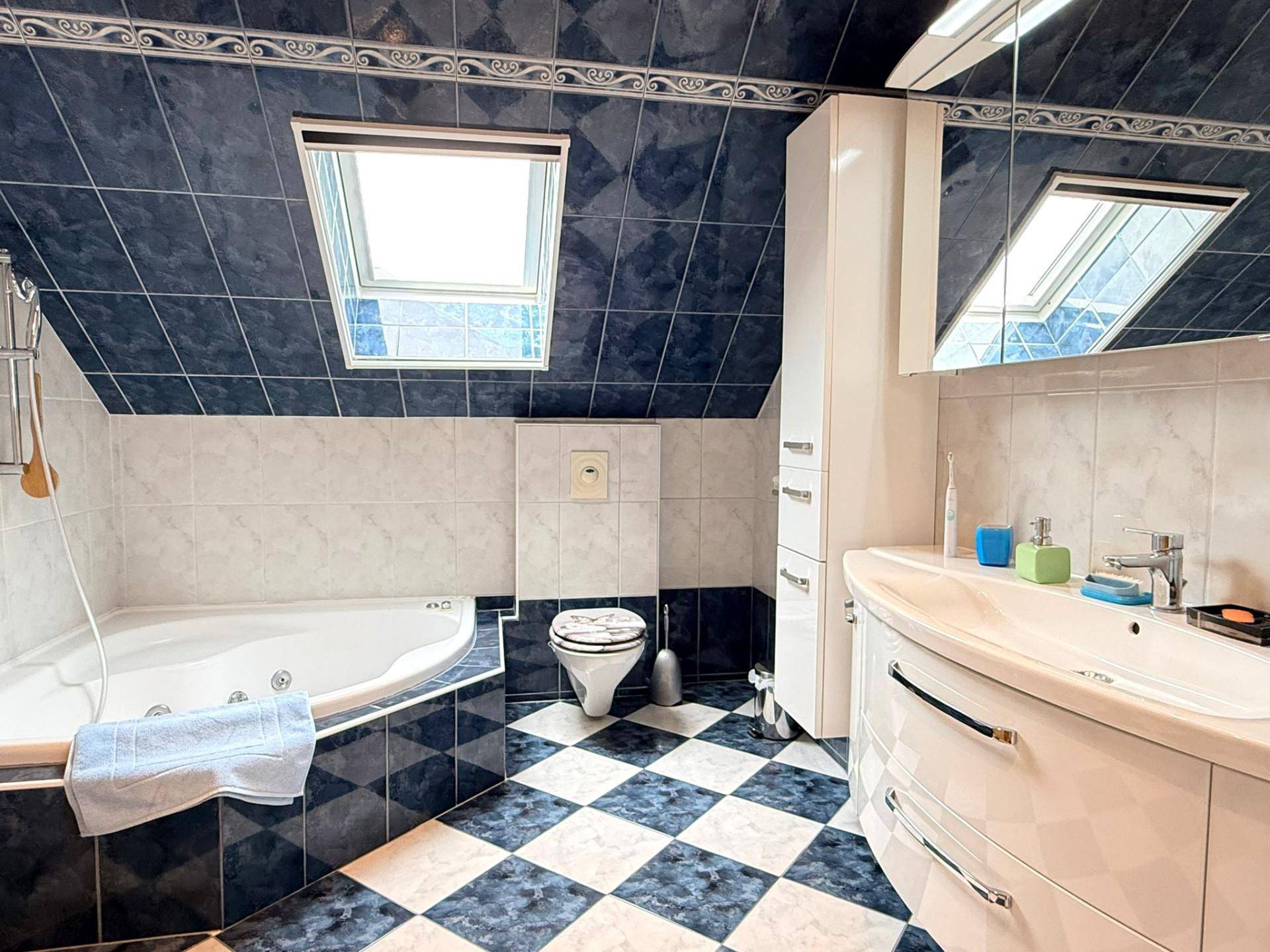 Modernes Badezimmer im Dachgeschoss mit Eckbadewanne, Dachfenster und blau-weiß karierten Fliesen.