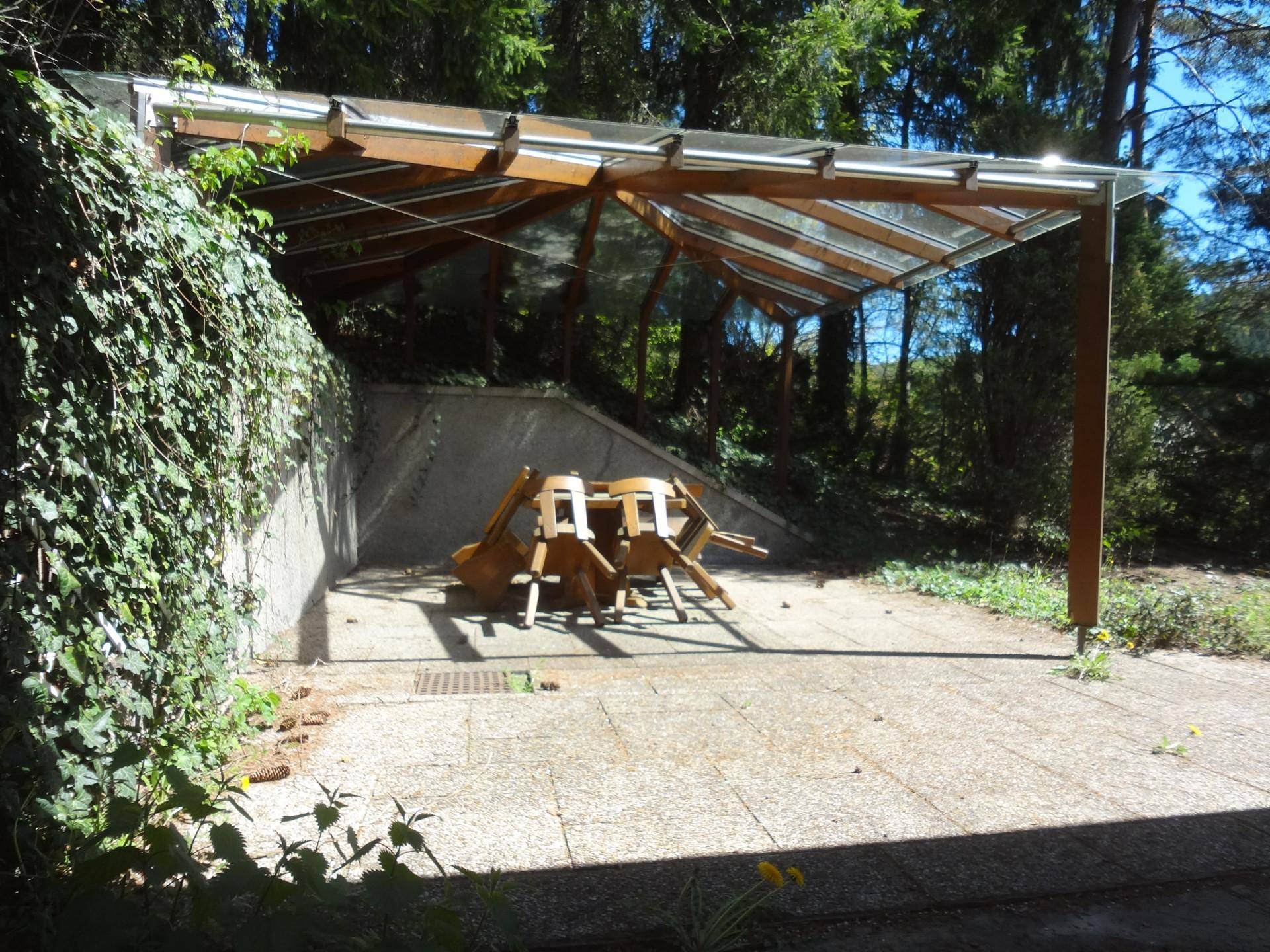 Überdachter Außenbereich mit Holz-Glas-Konstruktion und gepflastertem Boden, umgeben von Vegetation.