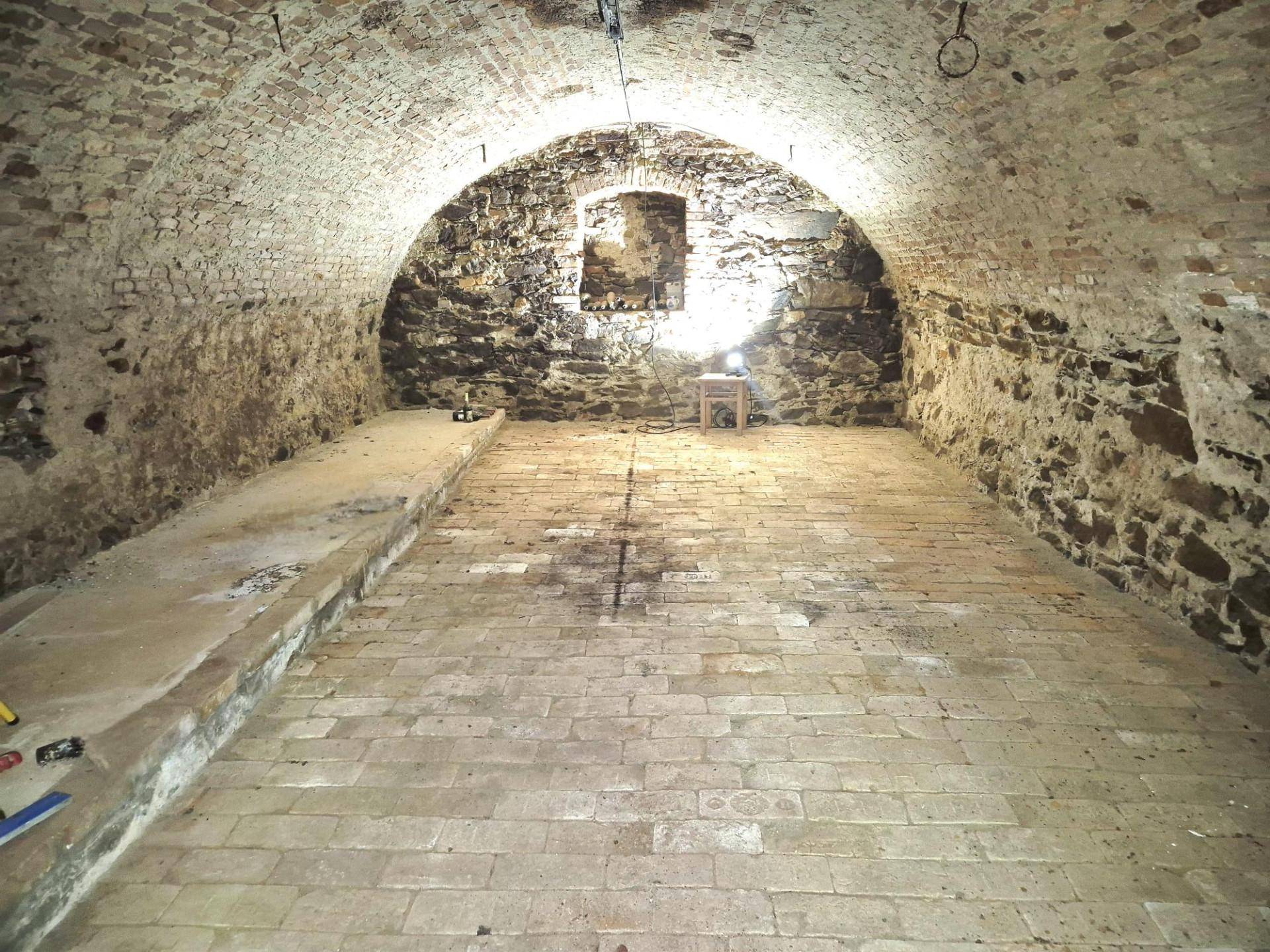Historischer Gewölbekeller mit Ziegelboden und freiliegenden Steinmauern, beleuchtet von einer Lampe.