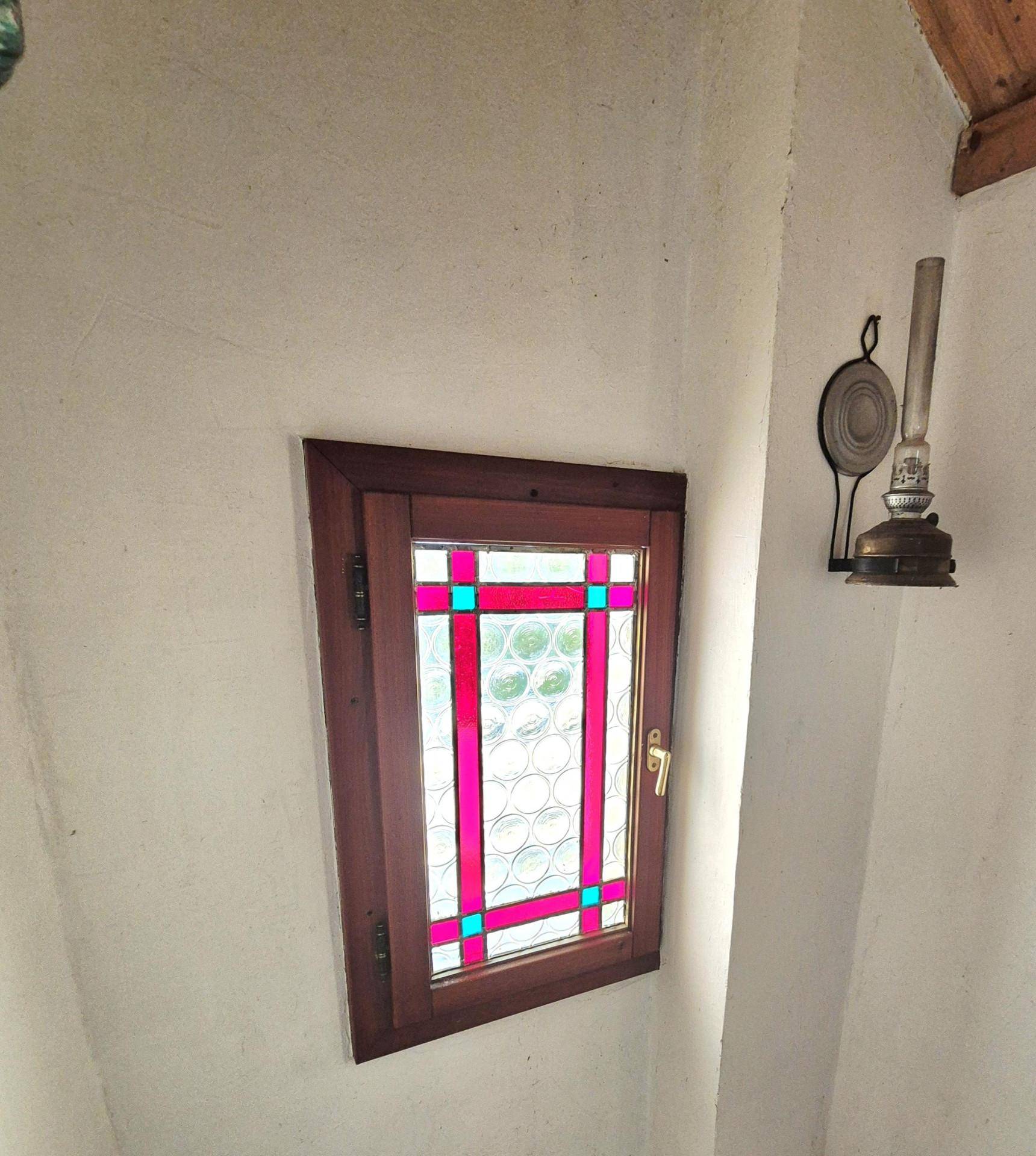 Kleines Fenster mit Buntglas und Holzrahmen in einer weiß verputzten Wand.