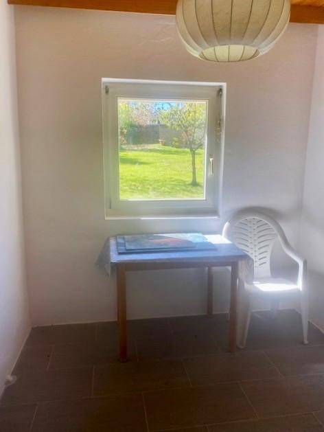Heller Raum mit Fenster zum Garten und einfacher Sitzgelegenheit.
