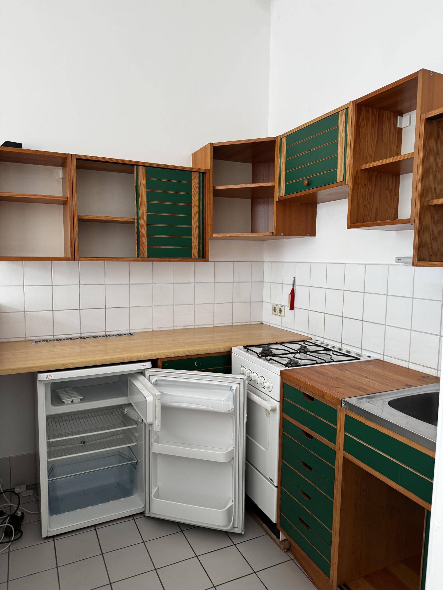 Ältere Küche mit grünen und holzfarbenen Schränken, Gasherd, Kühlschrank und Holz-Arbeitsplatte.