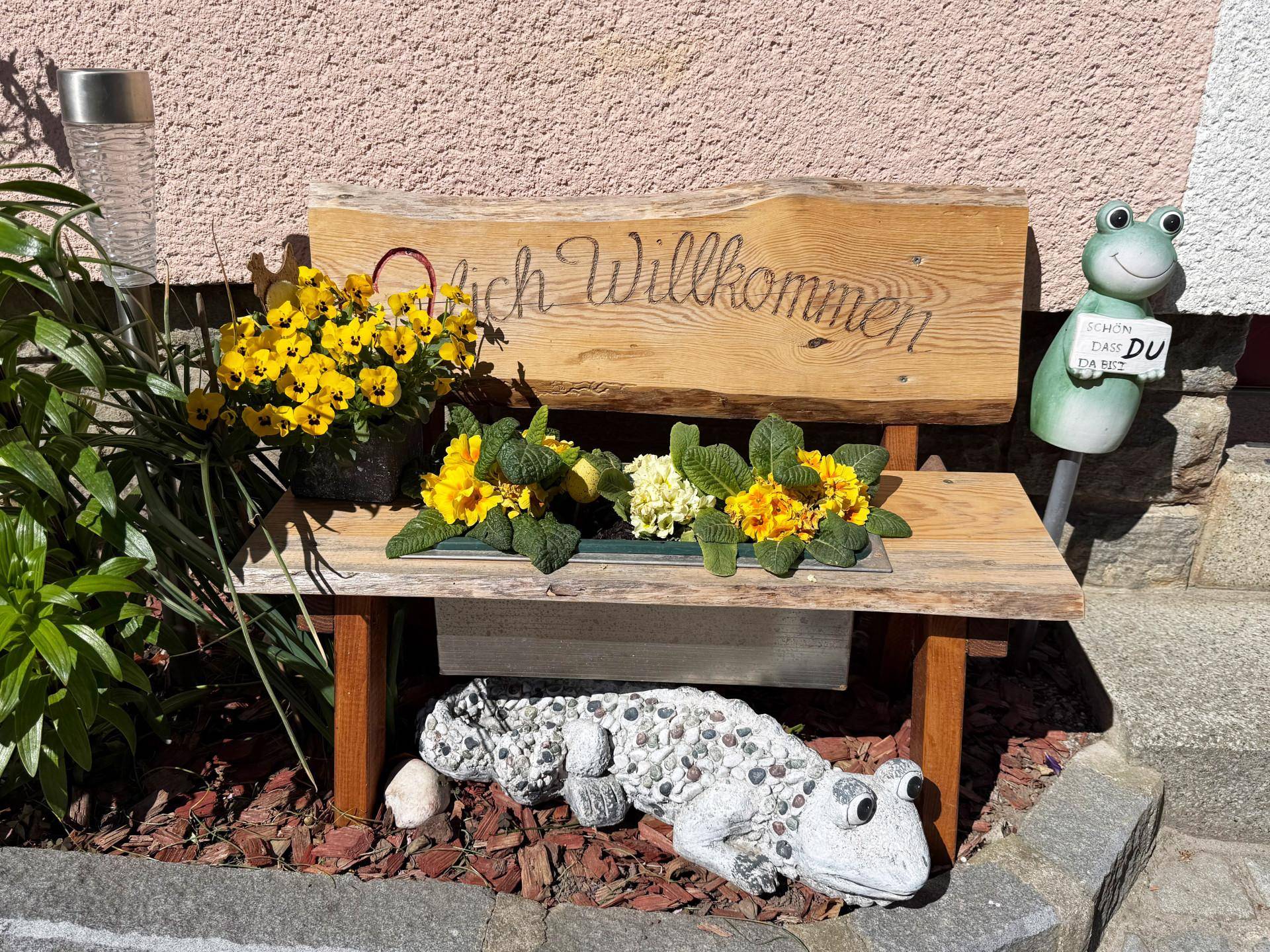 Eine kleine Holzbank mit Blumen und dekorativen Froschfiguren vor der Hauswand.