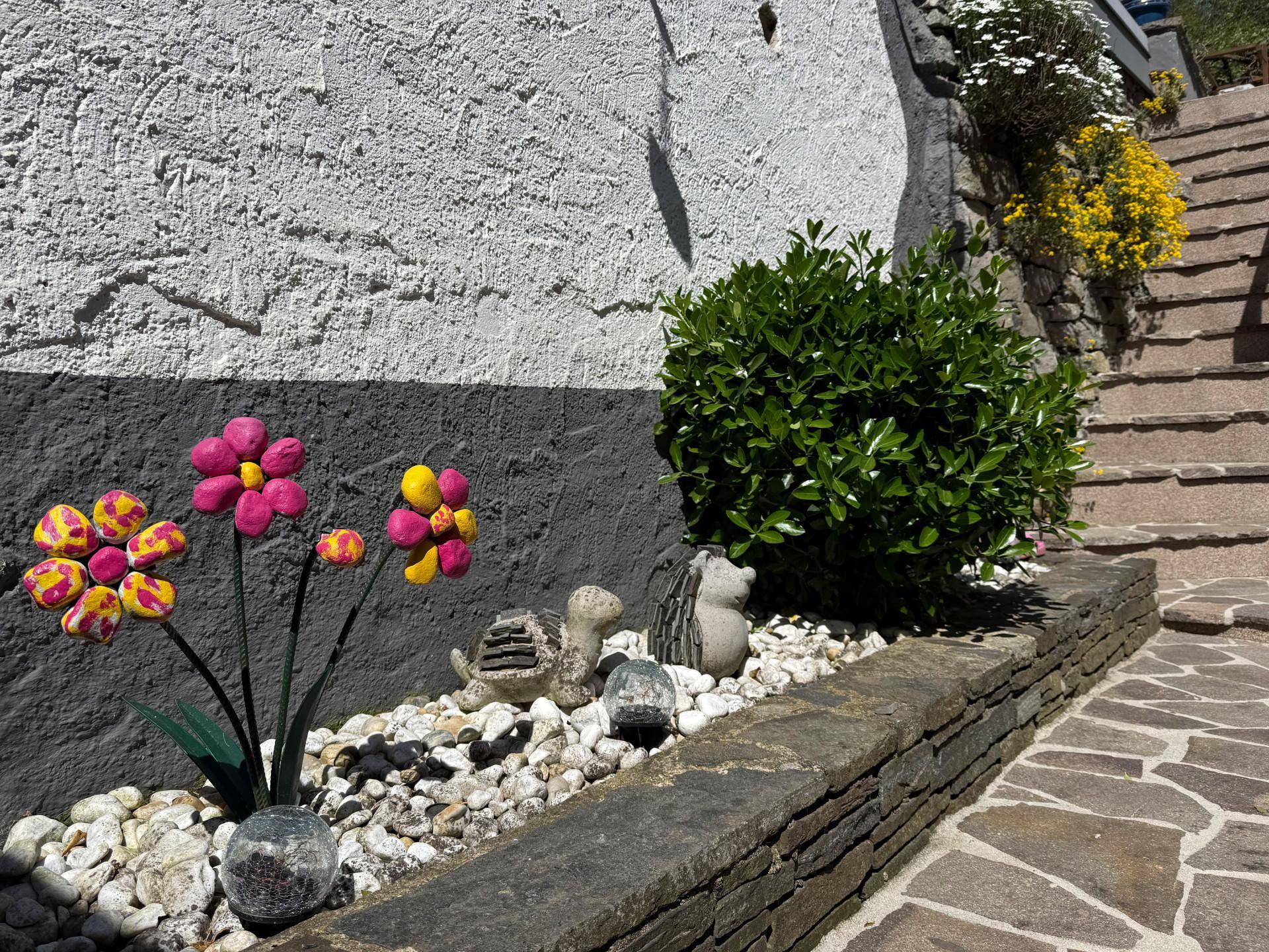 Dekorative Blumen und Steine entlang einer Hauswand mit einer Steintreppe im Hintergrund.