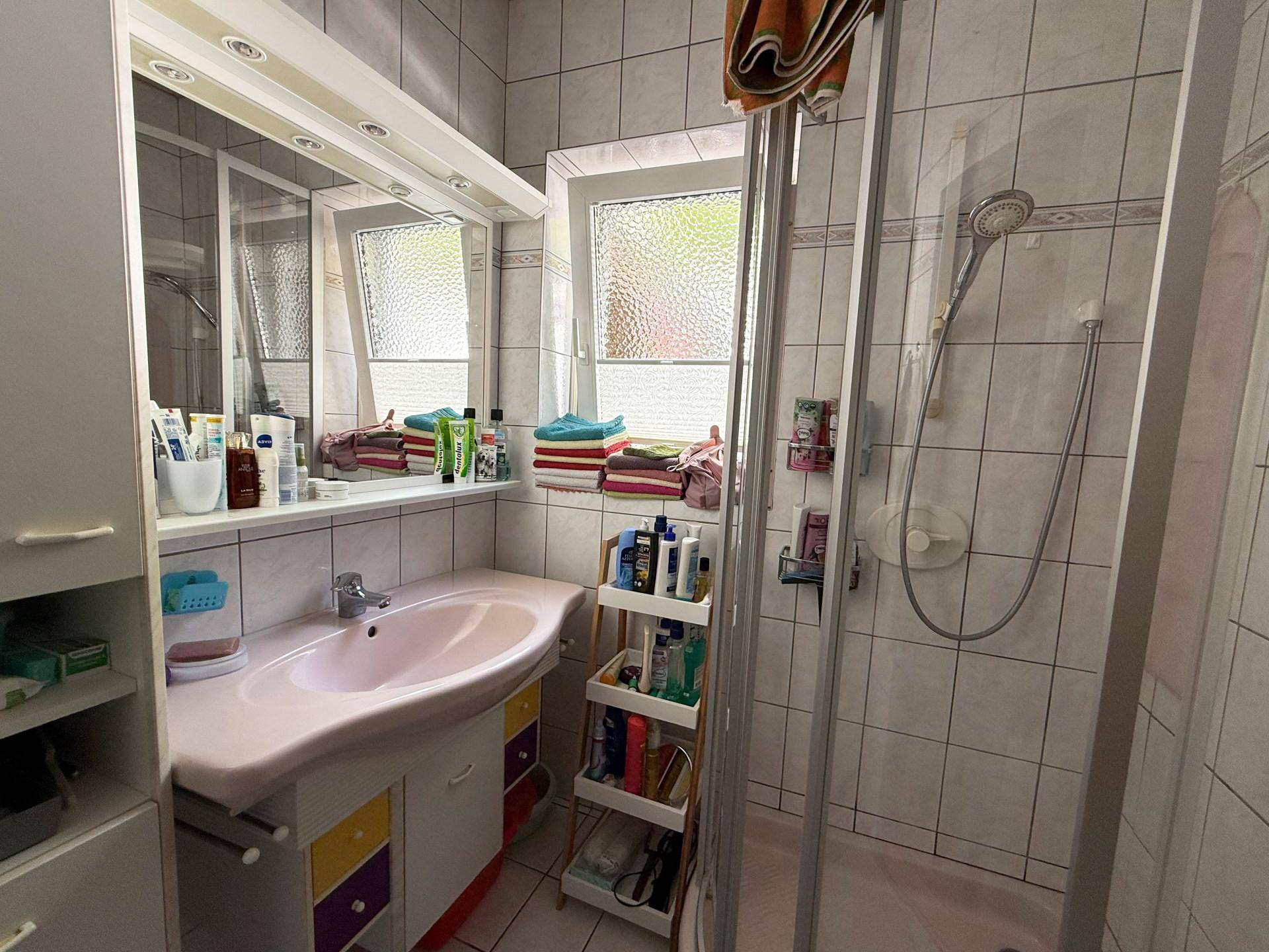 Badezimmer mit Dusche, Waschbecken und Fenster, ausgestattet mit hellen Fliesen.