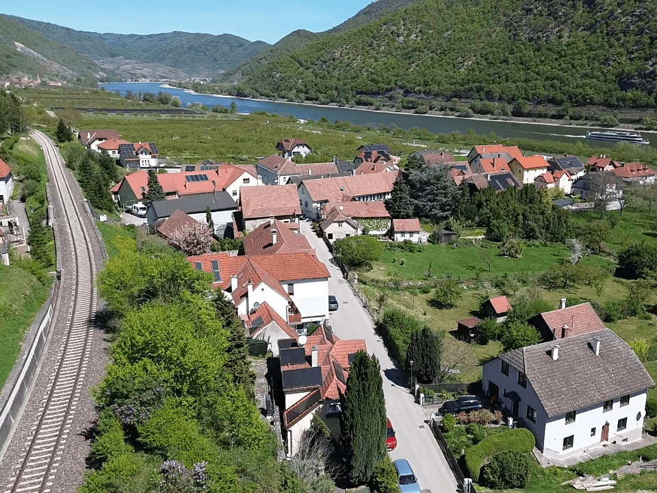Weitläufige Luftaufnahme eines Dorfes am Flussufer, umgeben von grünen Hügeln.