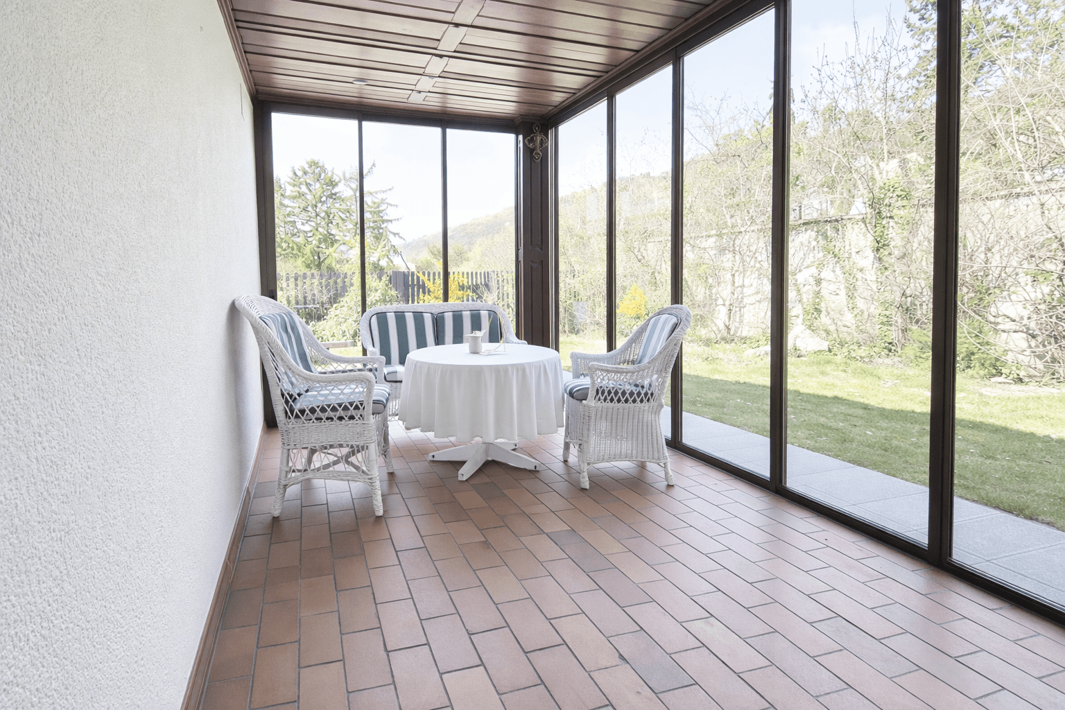 Gemütlicher Wintergarten mit Sitzgelegenheiten, Fliesenboden und weitem Blick ins Grüne.