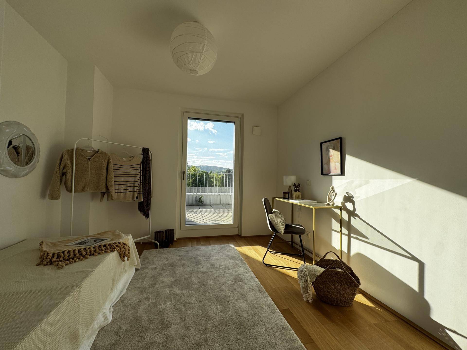 Schlafzimmer mit Holzboden, Schreibtisch und direktem Zugang zum sonnigen Balkon.