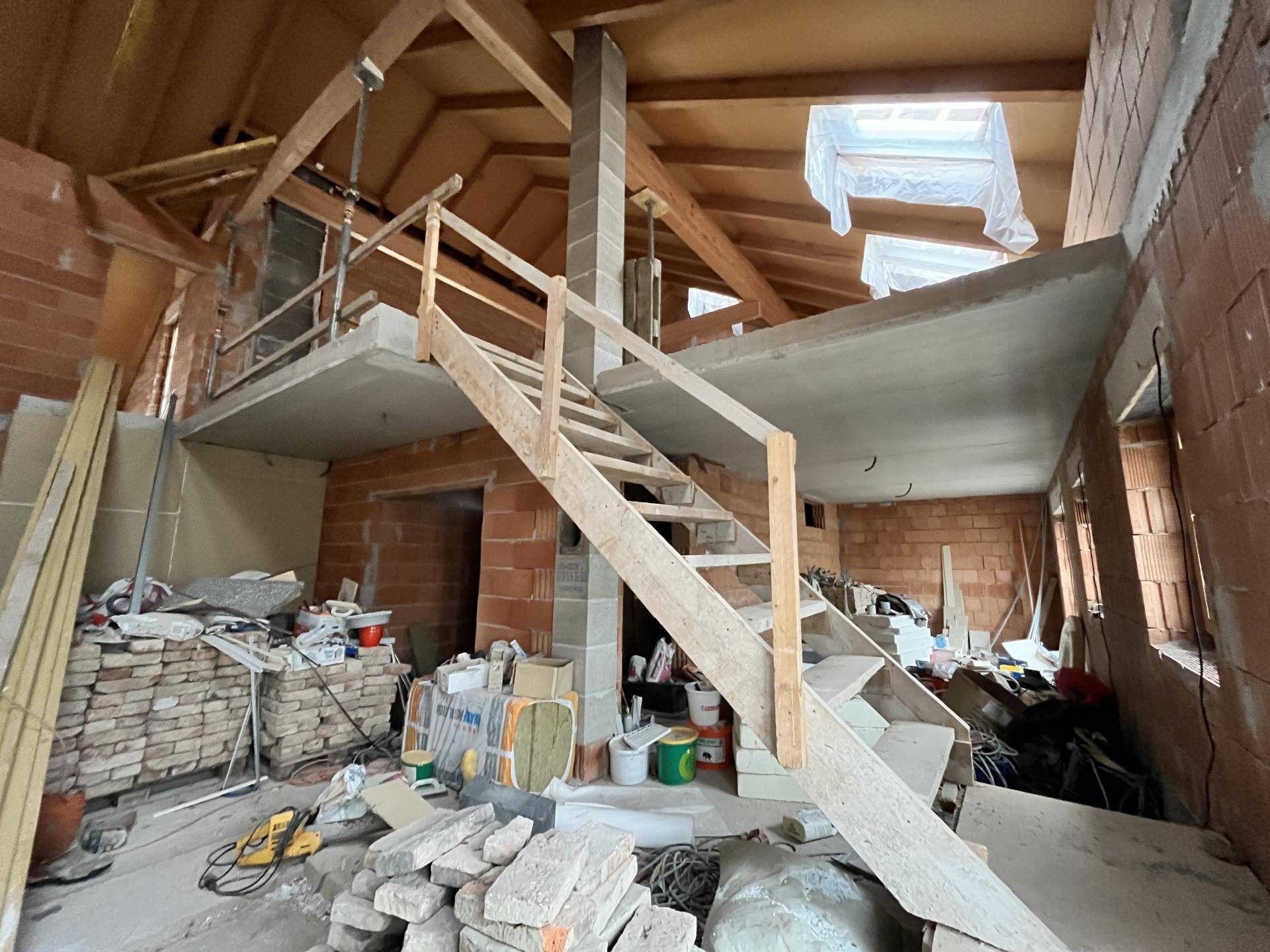 Innenraum im Rohbau mit Holzbalken, Treppe und Baumaterialien.