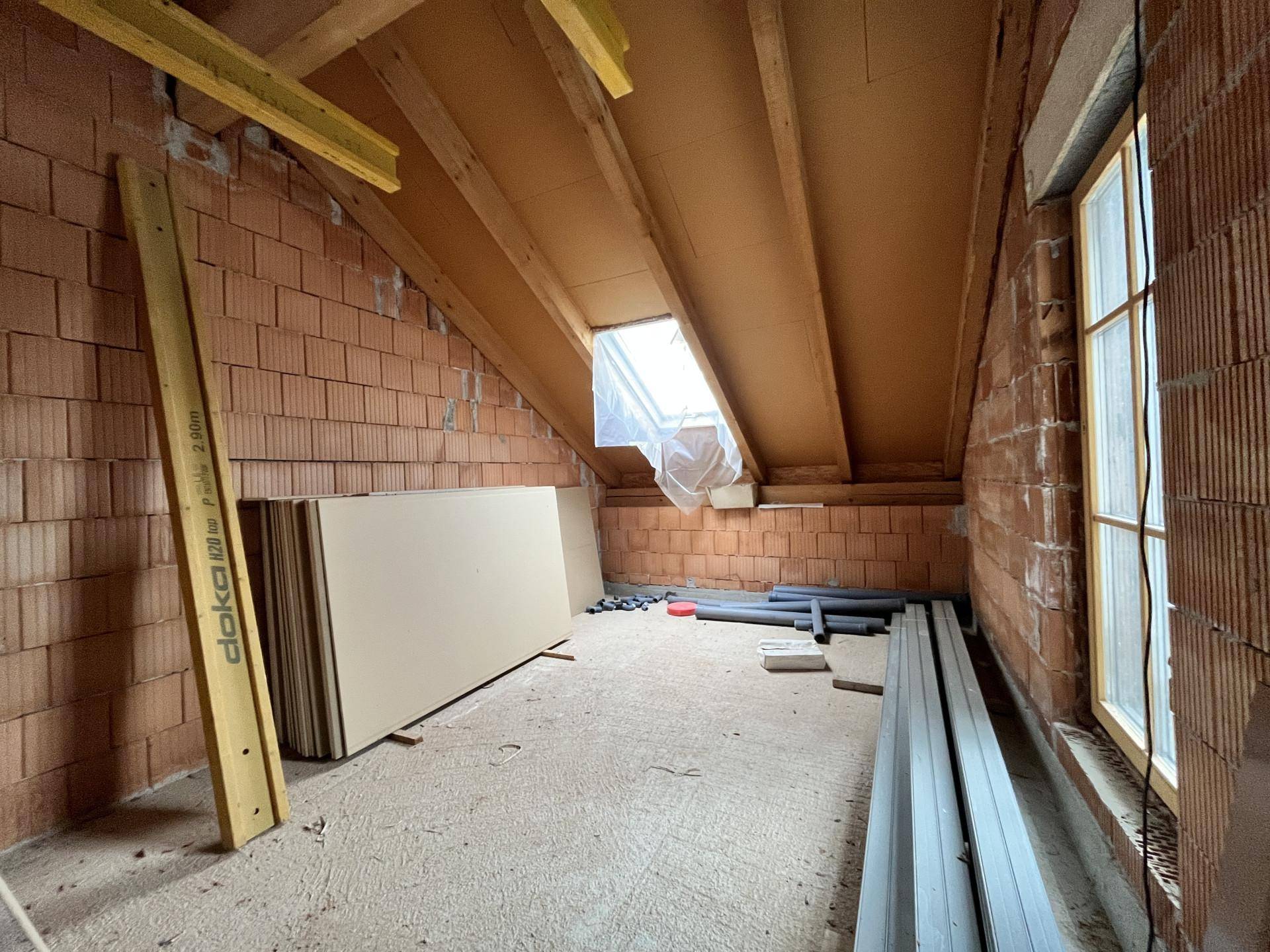 Dachgeschoss im Rohbau mit Holzbalken, Dachfenster und Ziegelwänden.