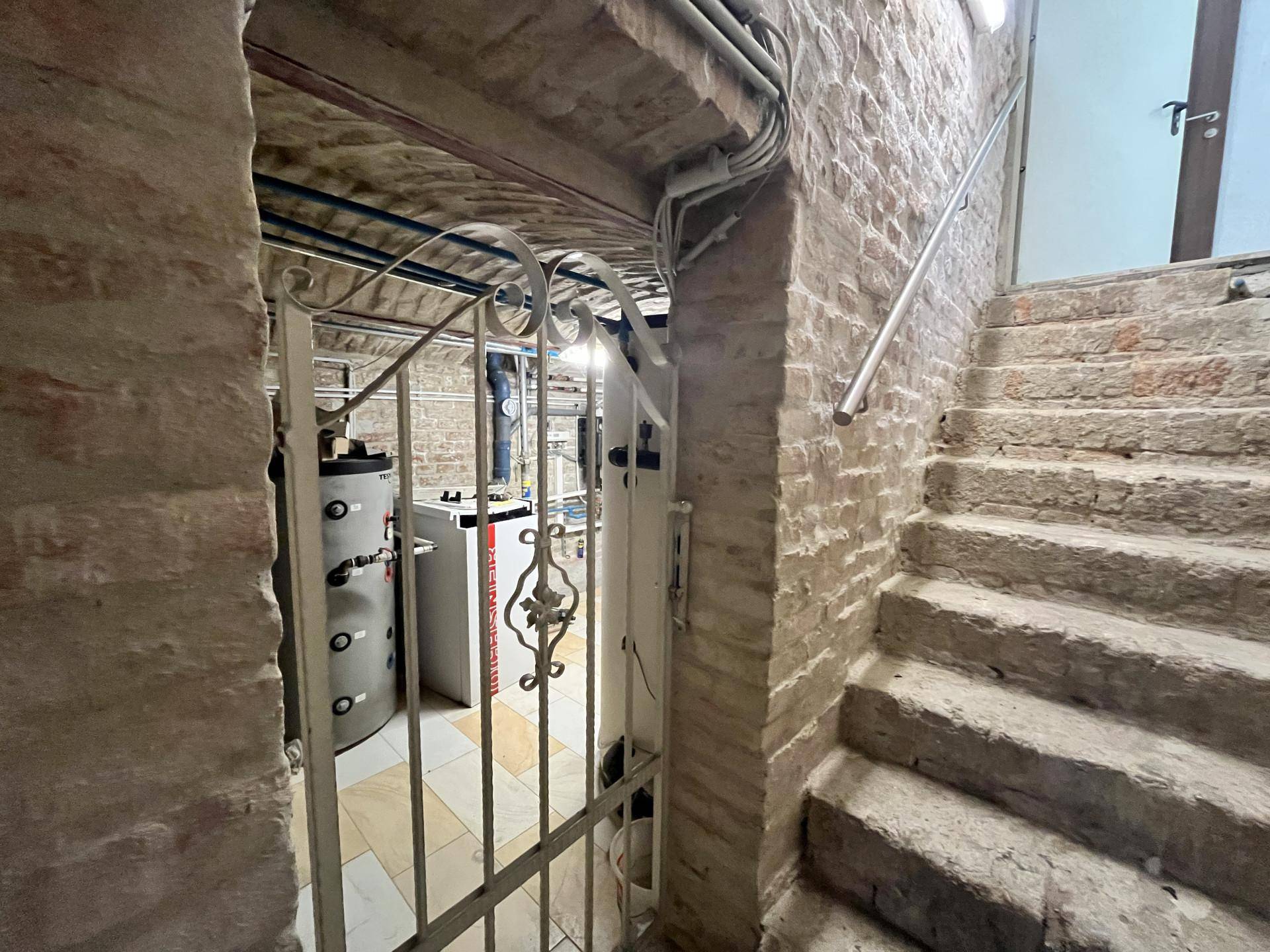 Kellerraum mit Ziegelwänden, Steintreppe und Heizungsanlagen, durch ein Gittertor zugänglich.