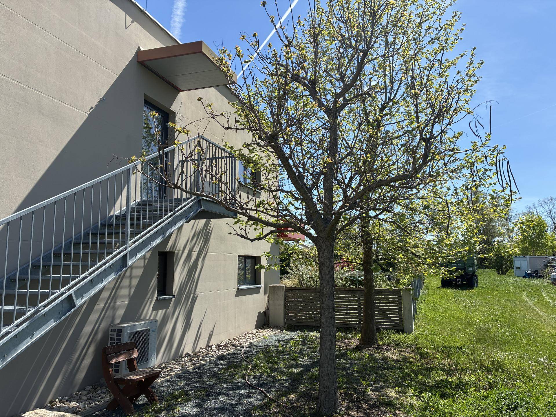Seitlicher Blick auf ein Gebäude mit Außentreppe aus Metall und einem Baum im Vordergrund unter klarem Himmel.