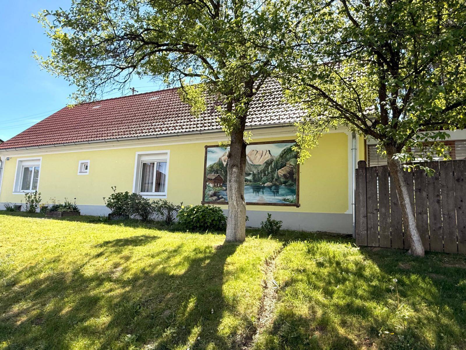 Gelbes Haus mit rotem Ziegeldach, umgeben von Bäumen und einem gemalten Wandbild.