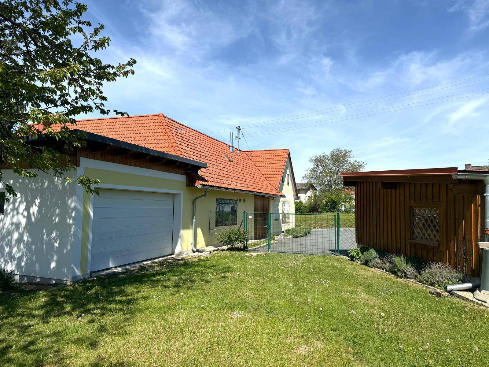 Gelbes Haus mit Garage und Holzschuppen, umgeben von einem grünen Rasen unter blauem Himmel.