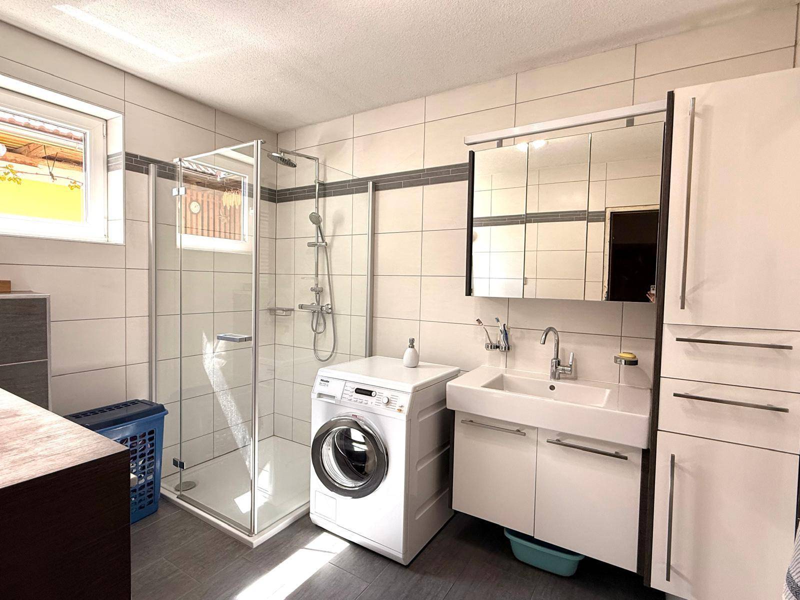 Modernes Badezimmer mit Duschkabine, Waschbecken und Waschmaschine, hell gefliest.