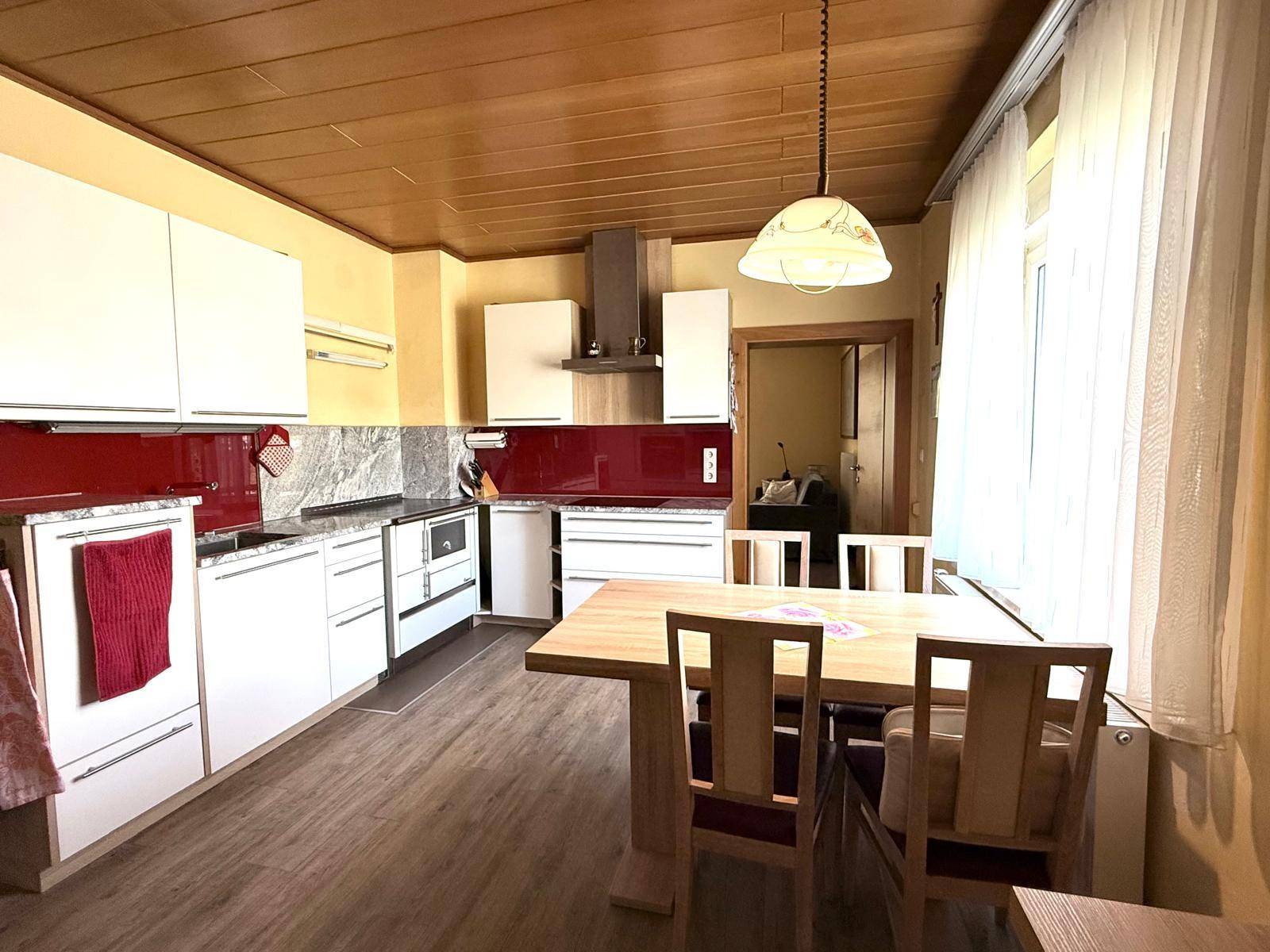 Moderne Wohnküche mit weißen Schränken, roter Rückwand und Esstisch unter Holzdecke.
