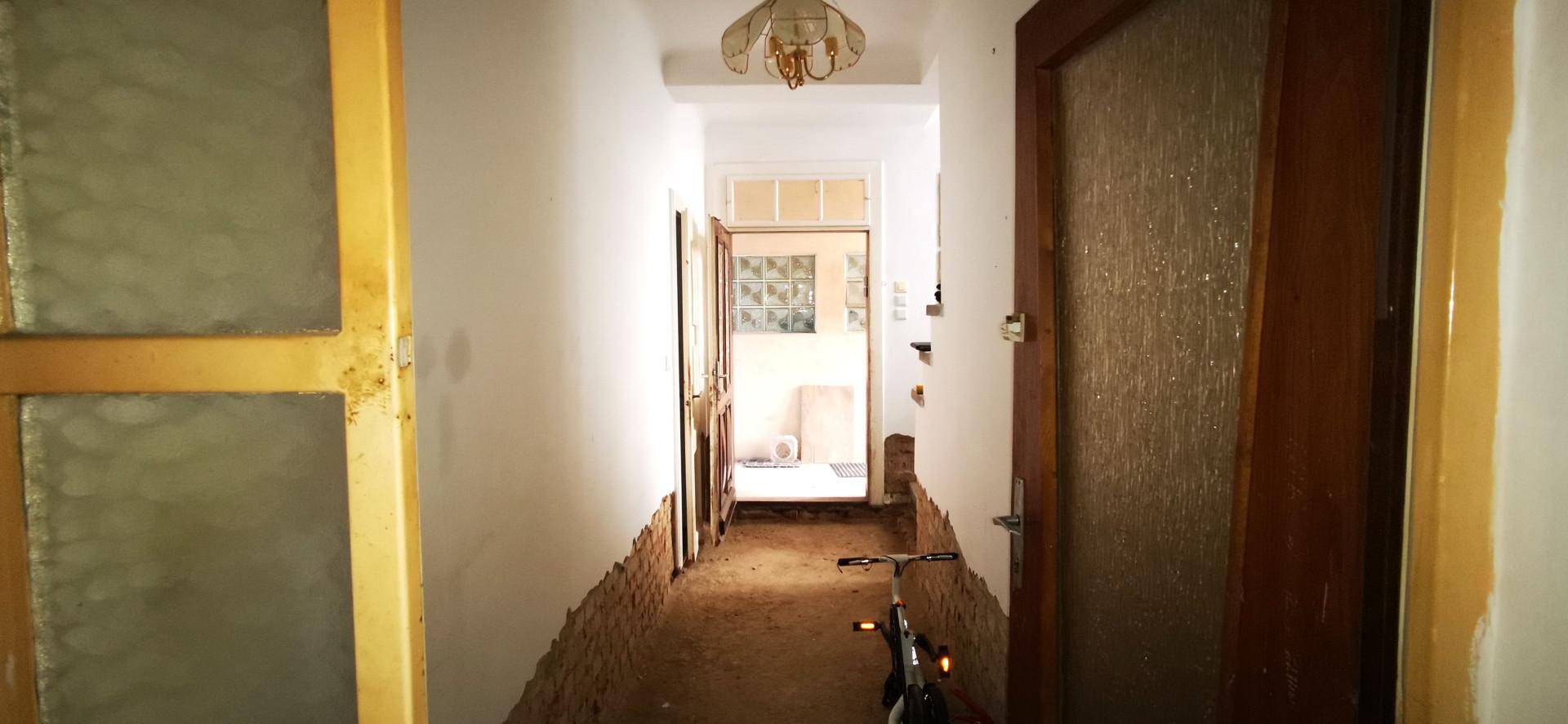 Langer Flur im Rohzustand mit freigelegten Ziegelwänden und alten Holztüren, bereit zur Renovierung.