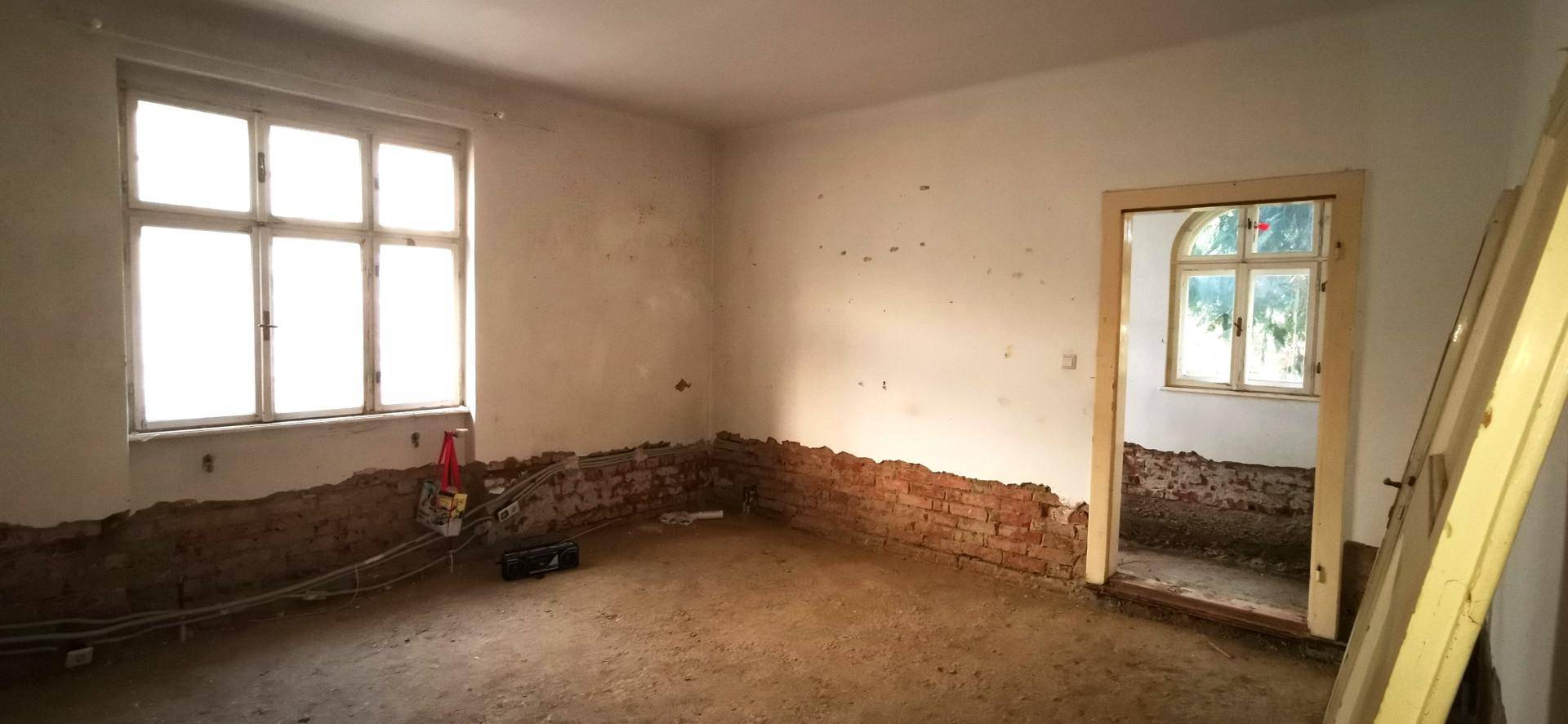 Renovierungsbedürftiger Raum mit freigelegten Ziegelwänden, großen Fenstern und Rohboden.