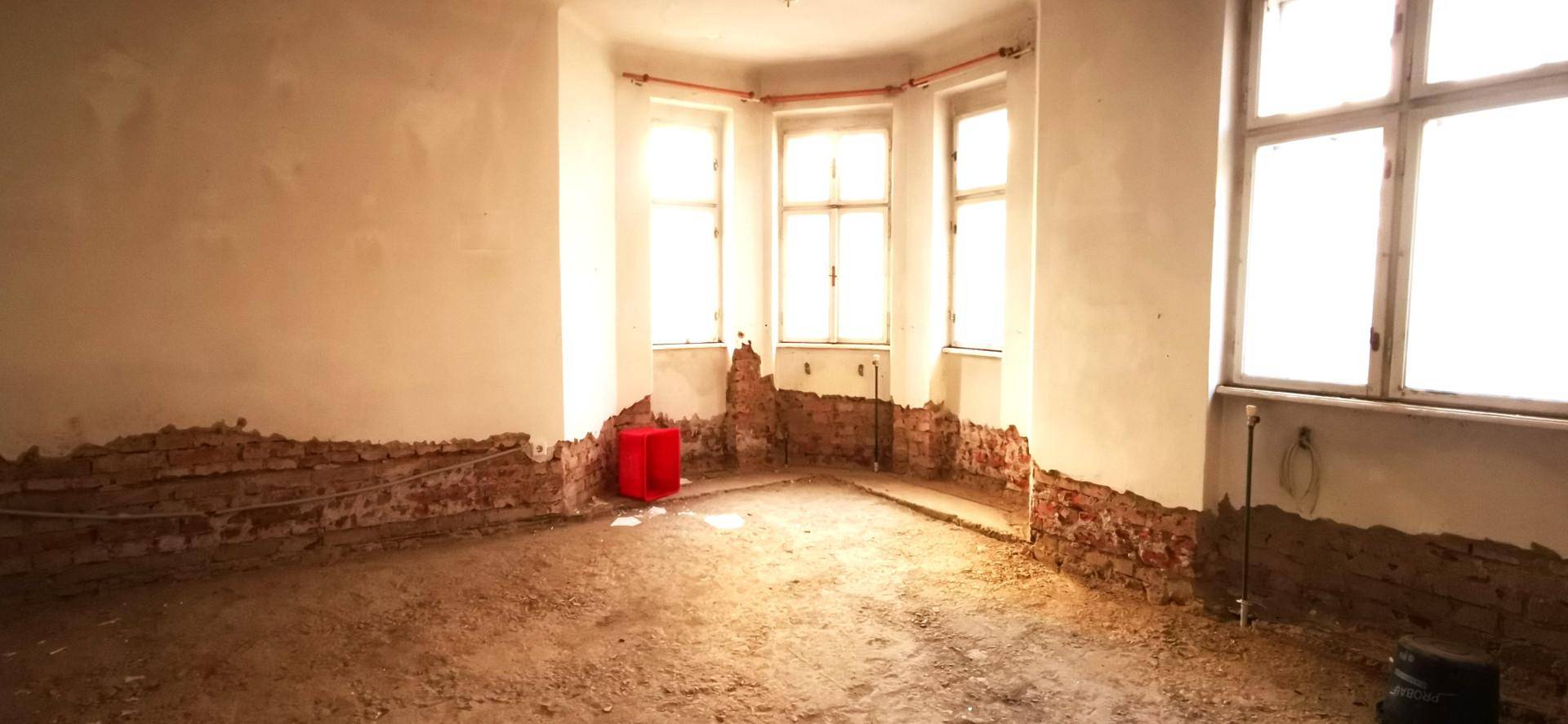 Eckraum im Rohbau mit Erkerfenstern, freigelegten Ziegeln und viel natürlichem Licht.