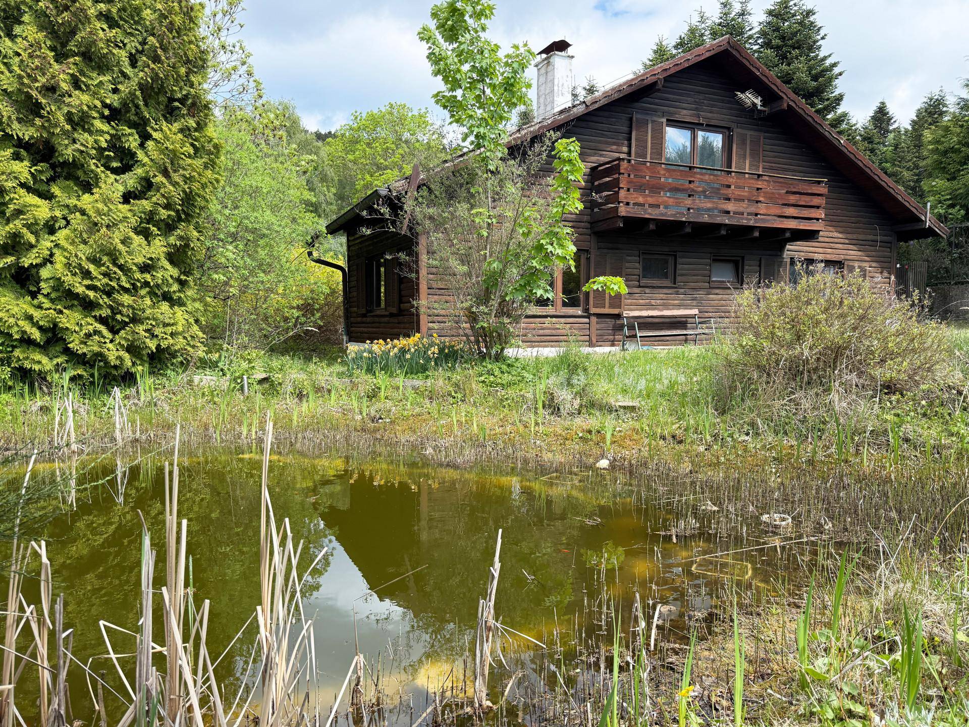 Charmantes Holzhaus mit Balkon, umgeben von üppigem Grün und einem natürlichen Gartenteich.