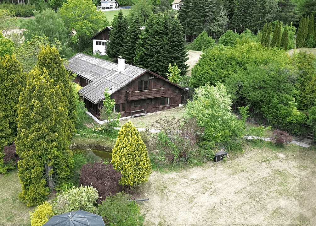 Luftaufnahme des Holzhauses eingebettet in eine grüne Gartenlandschaft mit Teich und hohen Bäumen.