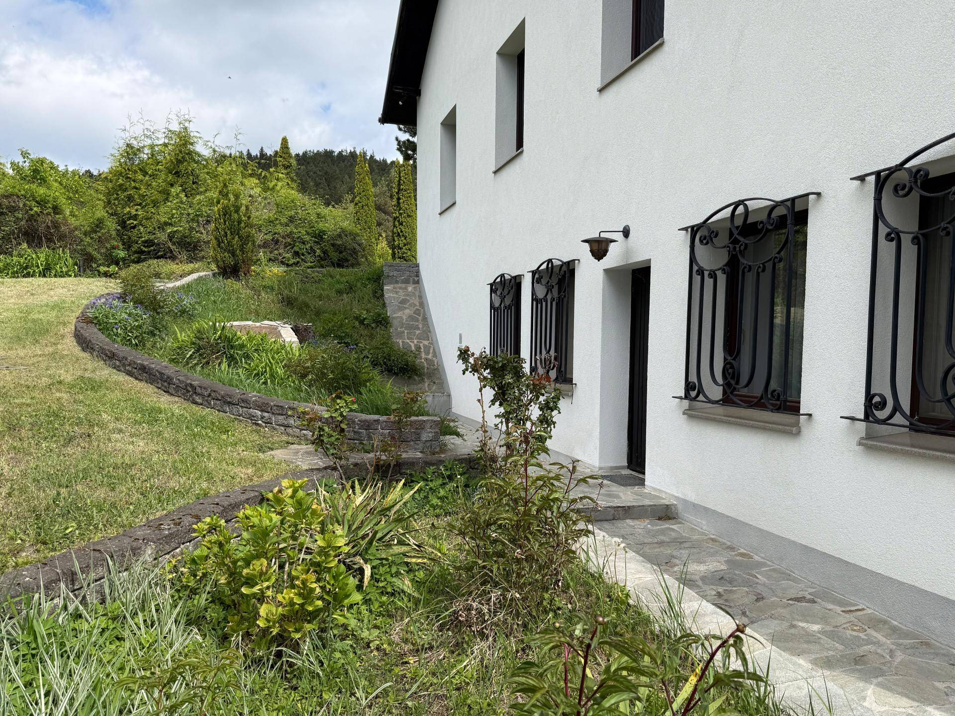 Seitenansicht des Haupthauses mit Natursteinmauer, gepflegter Bepflanzung und Zugang zum Garten.