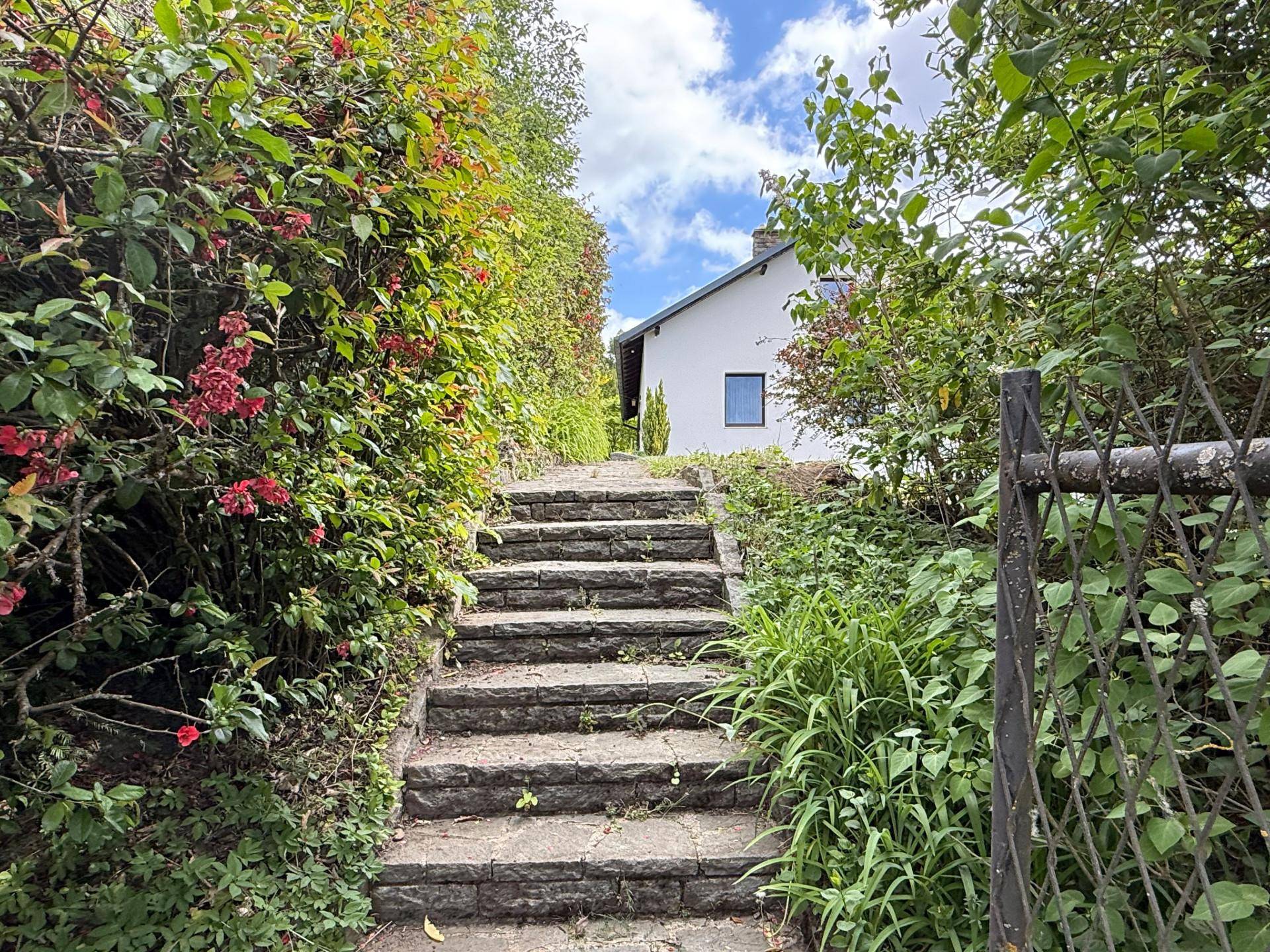 Natursteintreppe führt durch üppige Vegetation hinauf zum Haupthaus unter bewölktem Himmel.