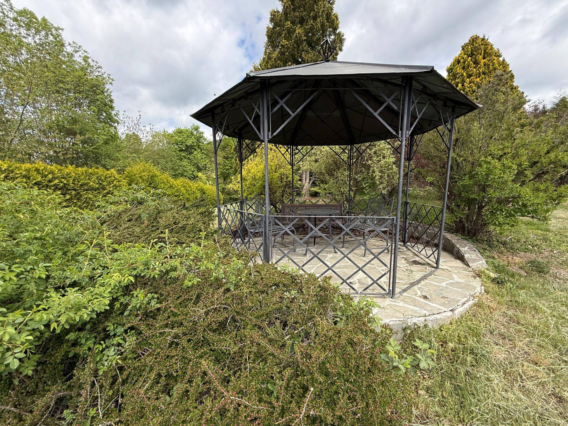 Detailansicht eines schmiedeeisernen Pavillons im Garten, umgeben von grünen Sträuchern und Bäumen.
