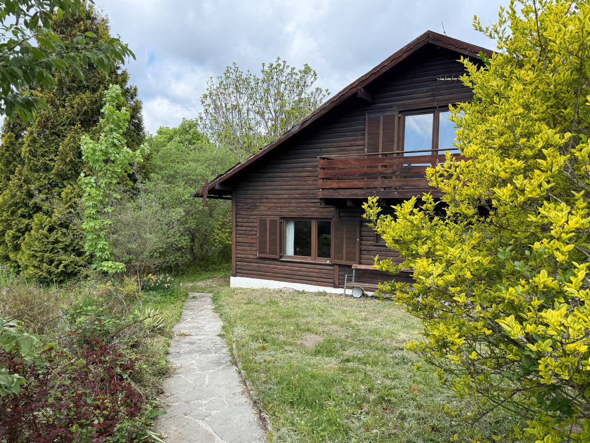 Rustikales Holzhaus mit Balkon, umgeben von einem grünen Garten mit Steinweg und Sträuchern.