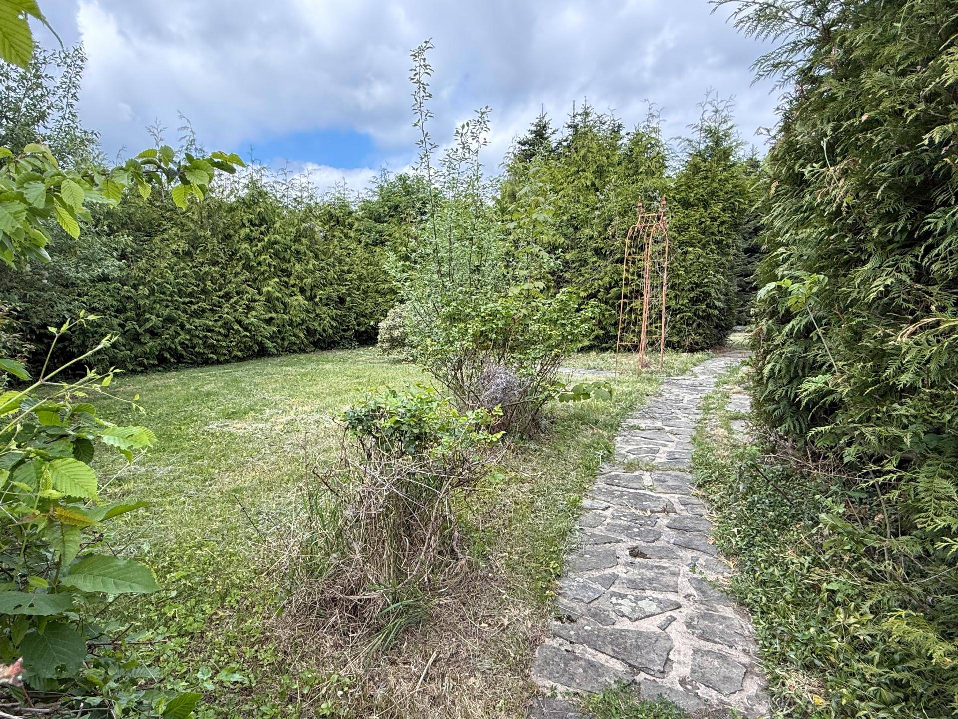 Steinweg führt durch einen gepflegten Garten mit Rasenflächen und hohen Hecken unter bewölktem Himmel.