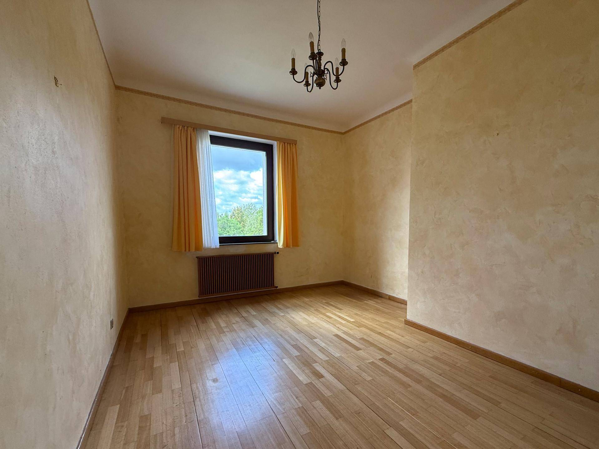 Helles Zimmer mit Holzparkettboden, gelben Wänden und einem Fenster mit Blick ins Grüne.