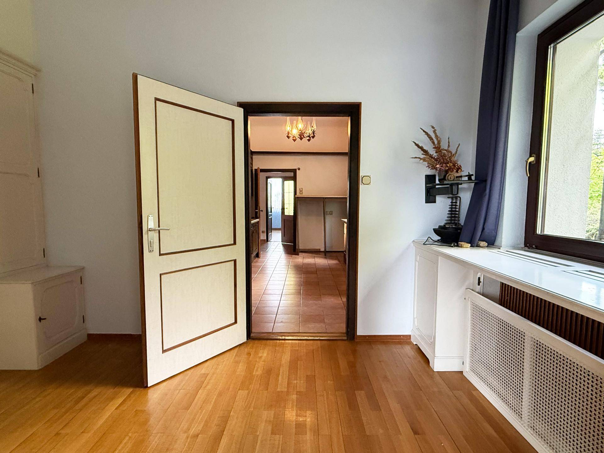 Helles Zimmer mit Holzparkettboden und Blick durch eine offene Tür in einen gefliesten Bereich.