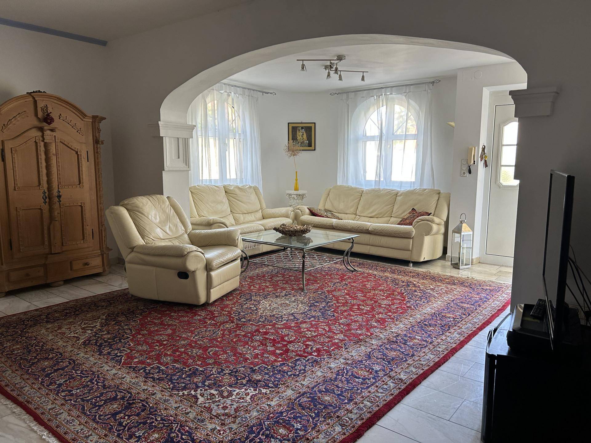 Geräumiges Wohnzimmer mit beigen Ledersofas, großem Teppich und Rundbogenfenstern.