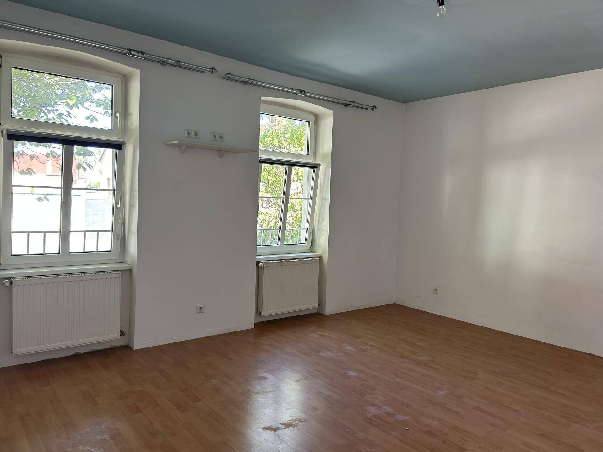 Geräumiges, leeres Wohnzimmer mit Holzboden, hohen Decken und großen Fenstern.