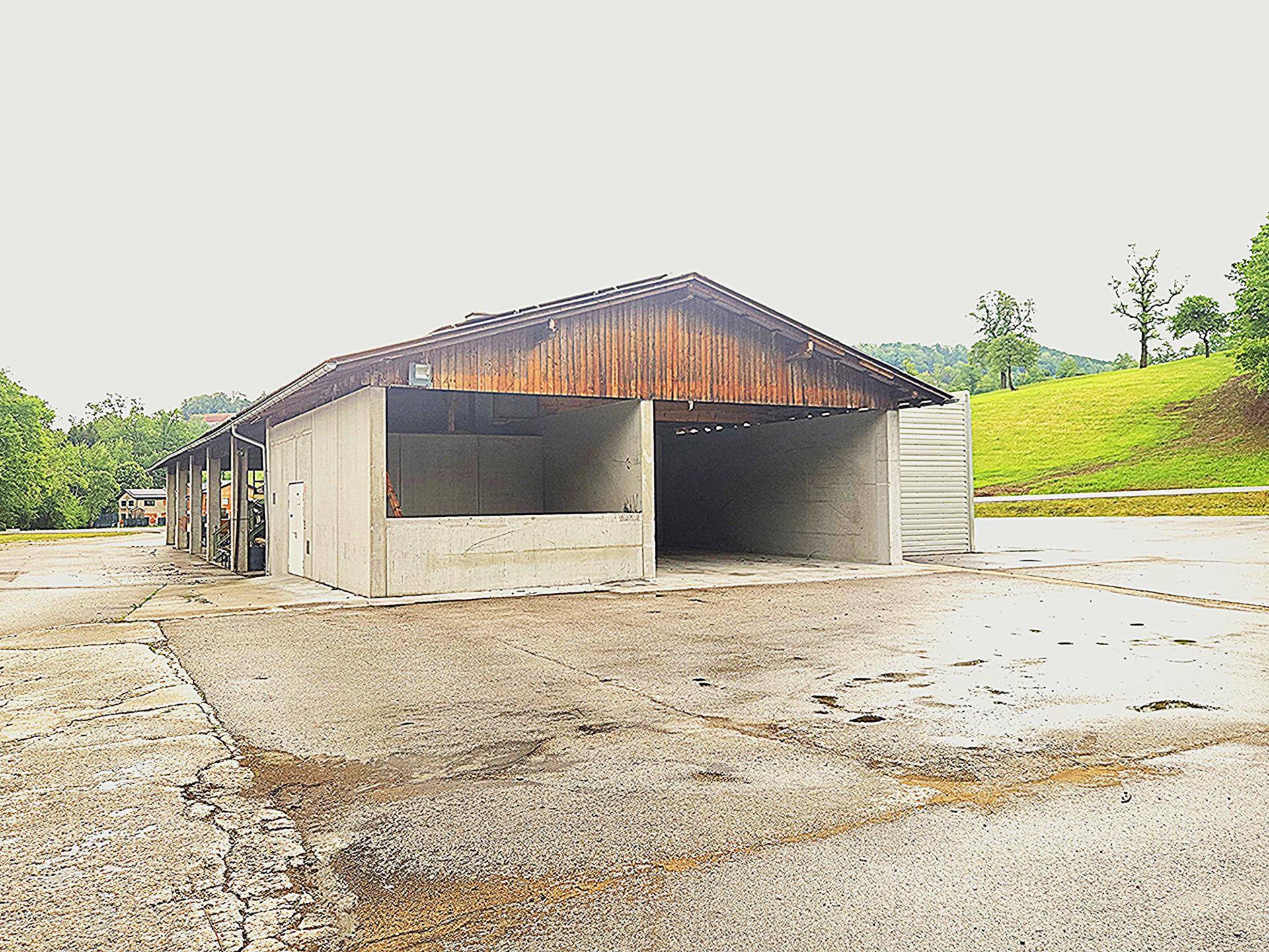 Offene Halle mit Holzdach und Betonwänden auf einer asphaltierten Fläche, umgeben von grünen Hügeln.